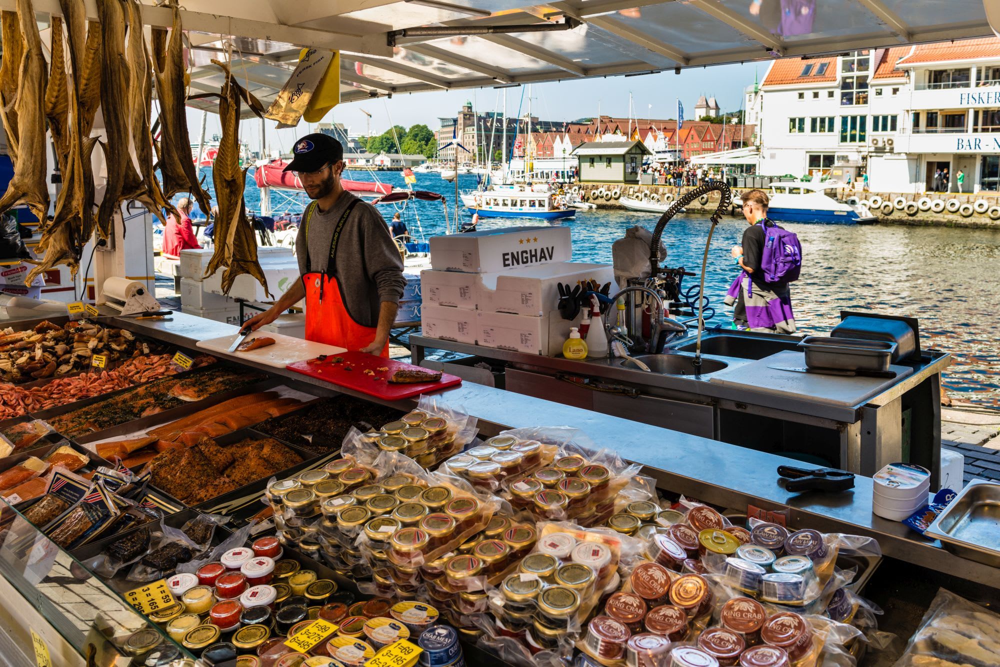 Fiskeselger på markedet i Bergen tilbereder sjømat omgitt av fisk, båter og bygg ved bryggen.