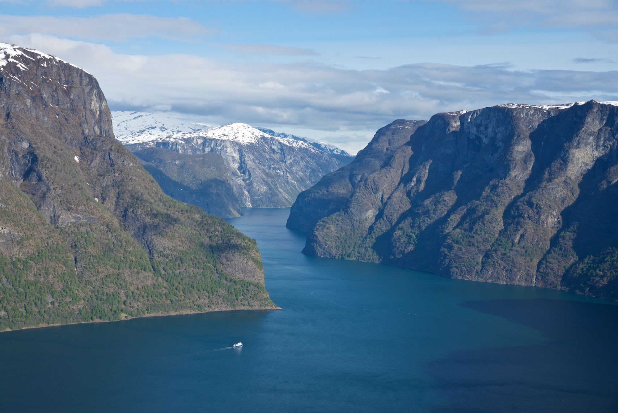 A white ferry sails through a calm fjord, flanked by snowy mountains under a clear sky.