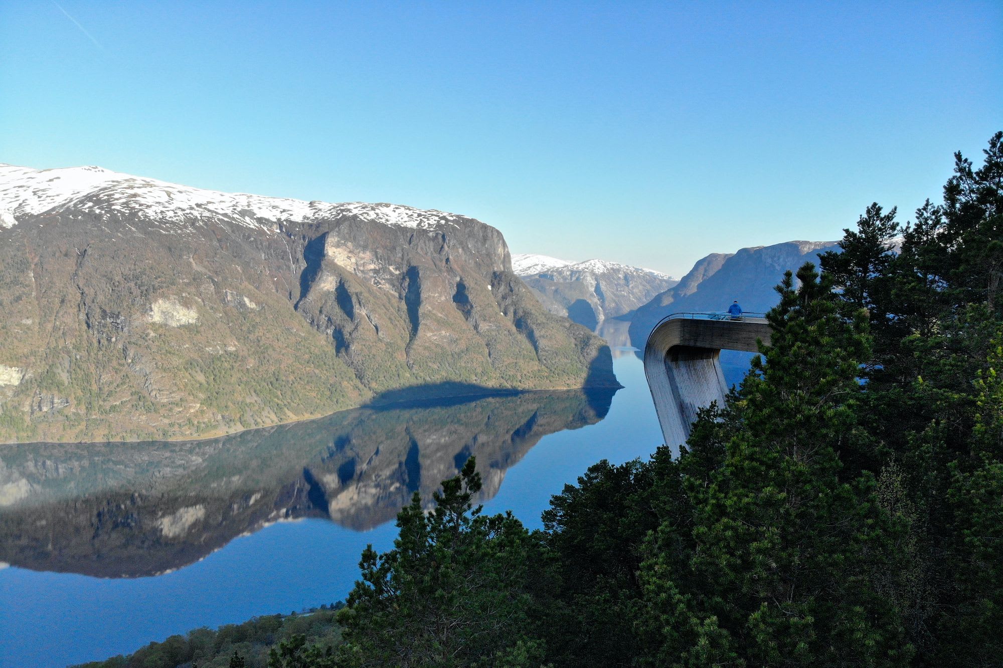 Person sits at end of curved viewing deck over fjord, with snowy peaks and clear blue sky.