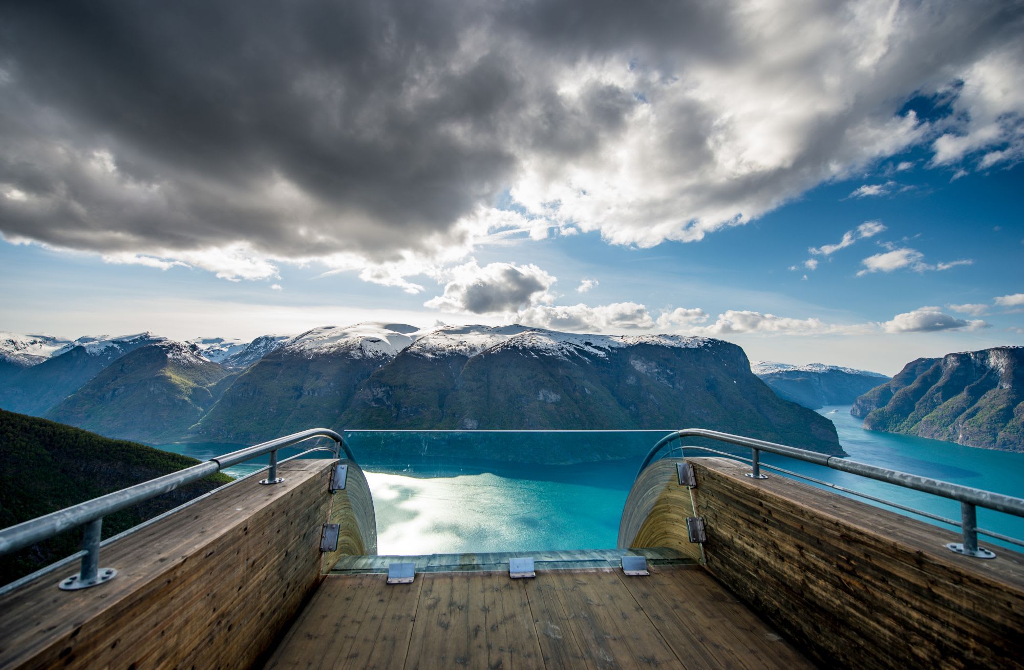 Viewpoint platform overlooking a dramatic fjord in Norway with turquoise water and snowy peaks under clouds.