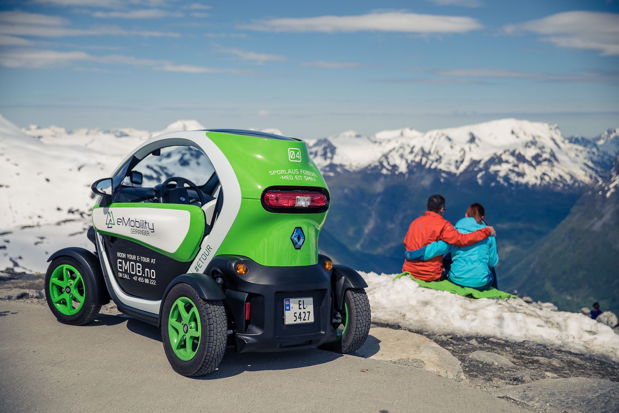 Electric tour car parked on snowy road in Geiranger with couple enjoying view of snowy peaks.