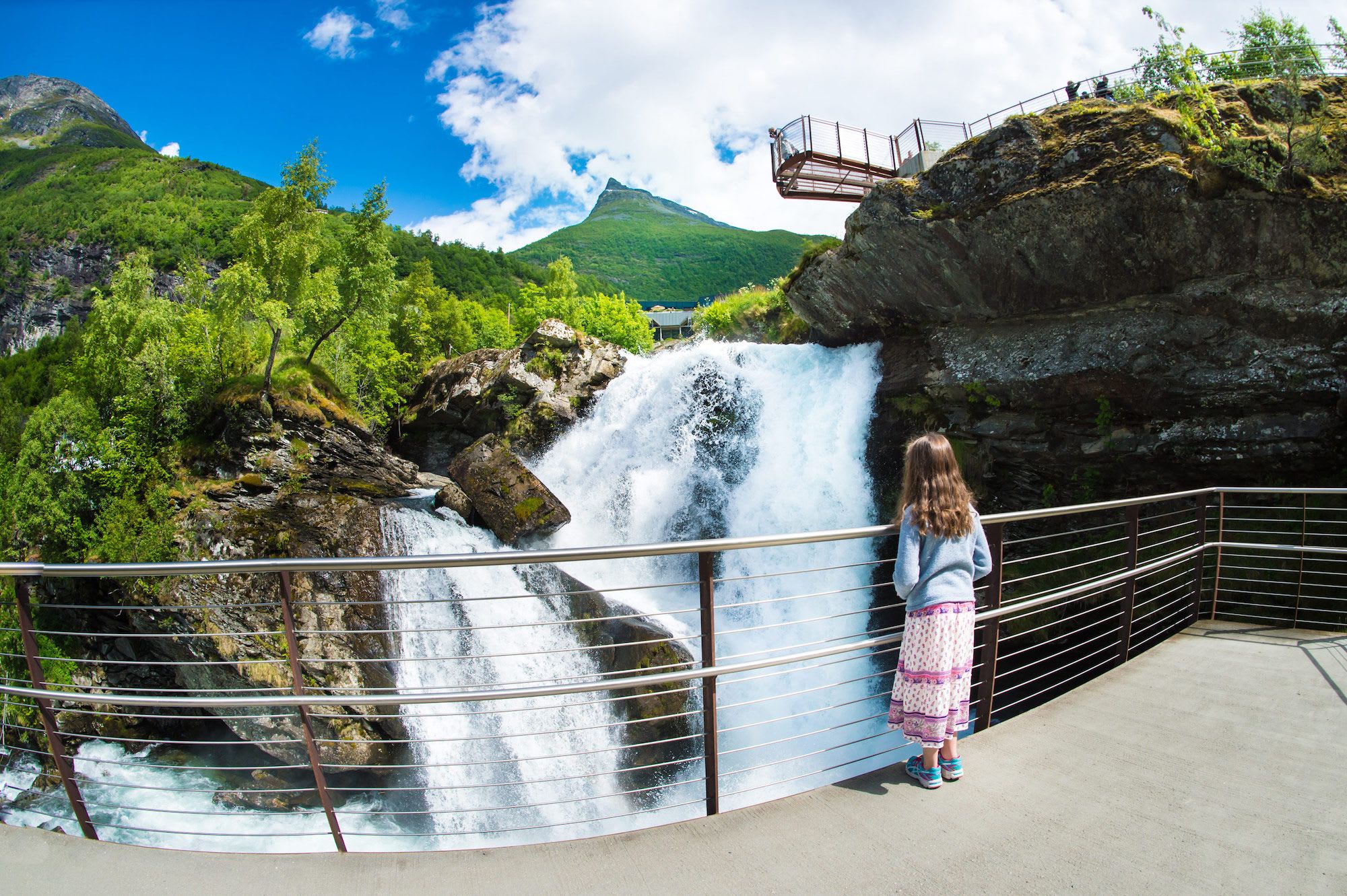 Girl stands on platform overlooking a waterfall and green mountains under a blue sky with scattered clouds.