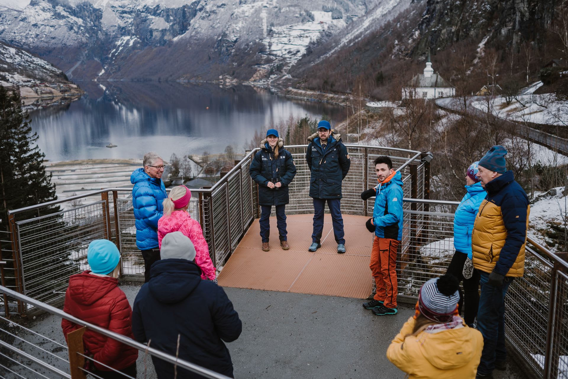 Group in winter clothes on platform overlooking calm fjord with snowy mountains and distant white church.