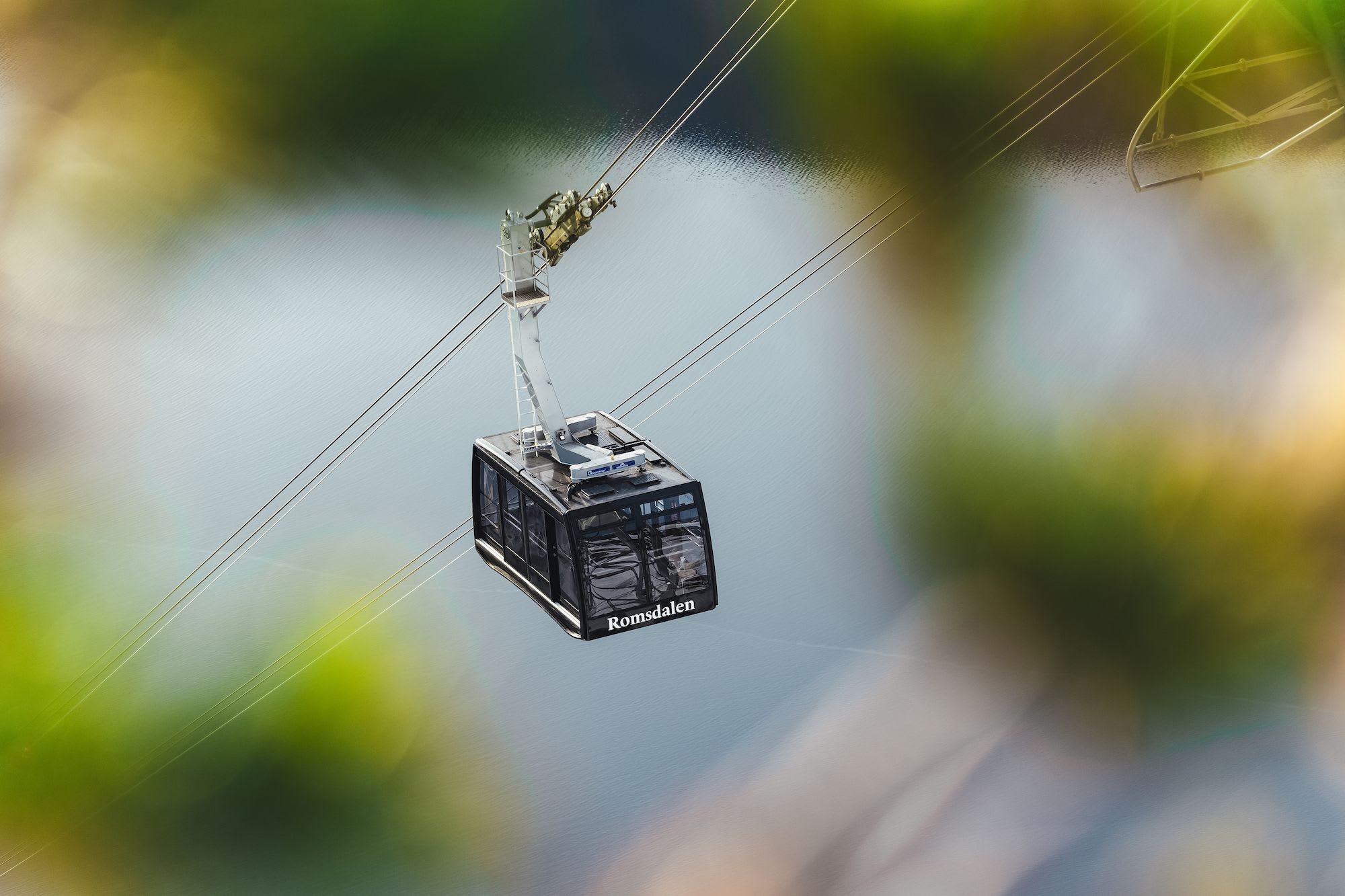 A close-up of a cable car with the "Romsdalen" logo, suspended above calm water with blurred landscape below.