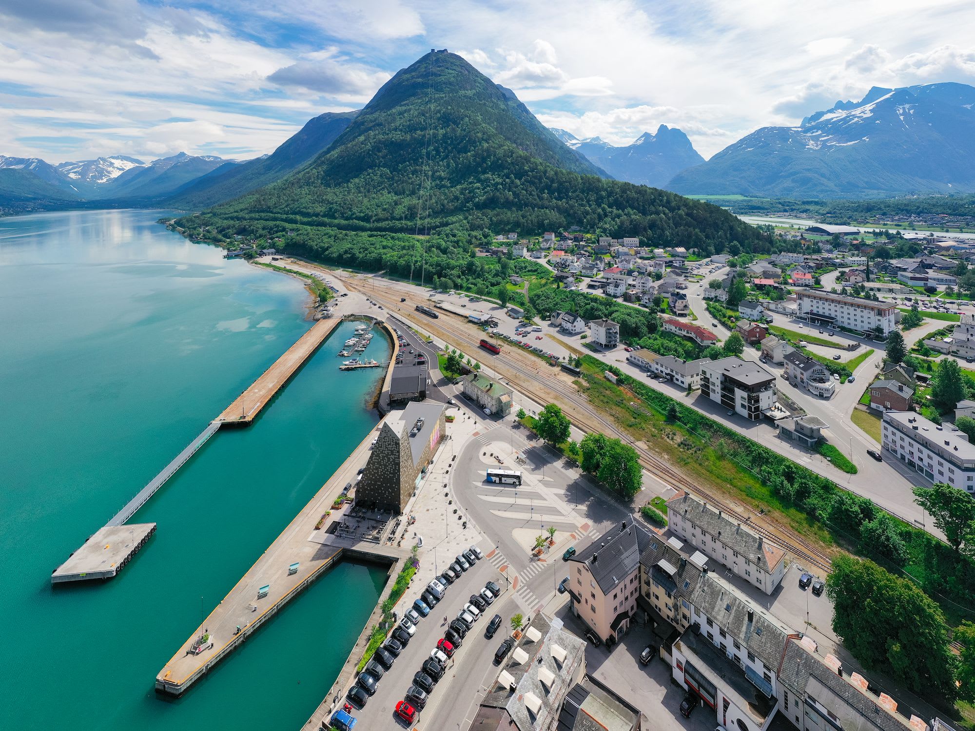 An aerial view of a scenic town by the water, with mountains towering in the distance and a pier stretching into the turquoise water.