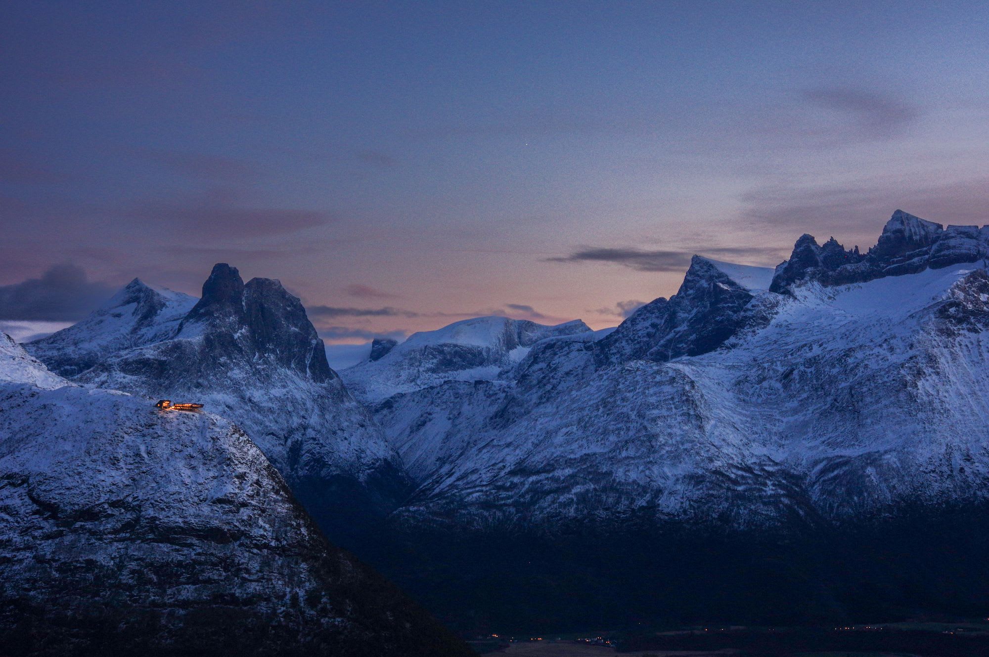 A stunning mountain landscape during dusk, with snow-covered peaks and a small building in the distance.