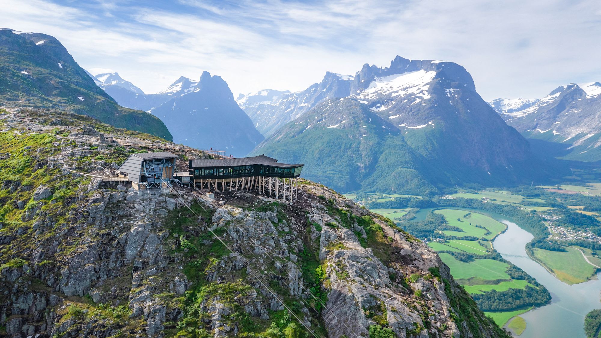 An aerial view of a modern building perched on a rocky outcrop, surrounded by snow-capped mountains.