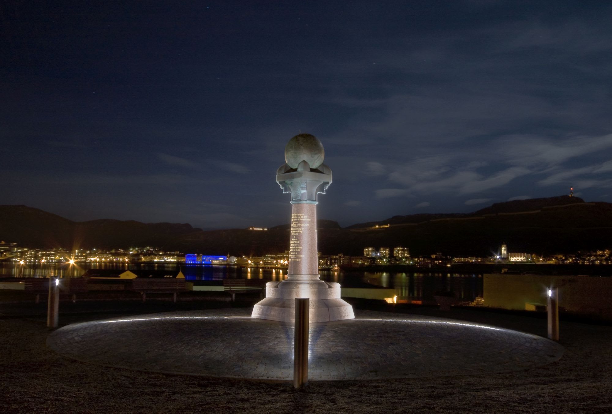 Høyt monument med globus på toppen lyser om natten, med bylys reflektert i vann.
