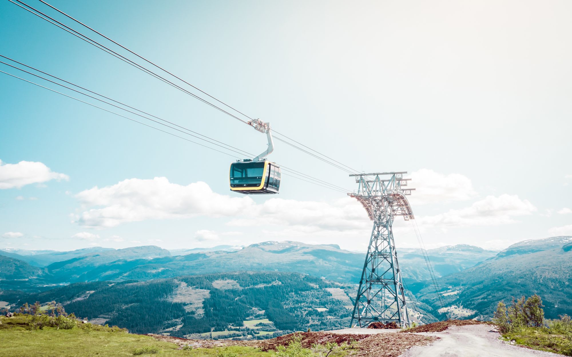 Yellow-black cable car travels high above green valleys and peaks under bright clear sky.