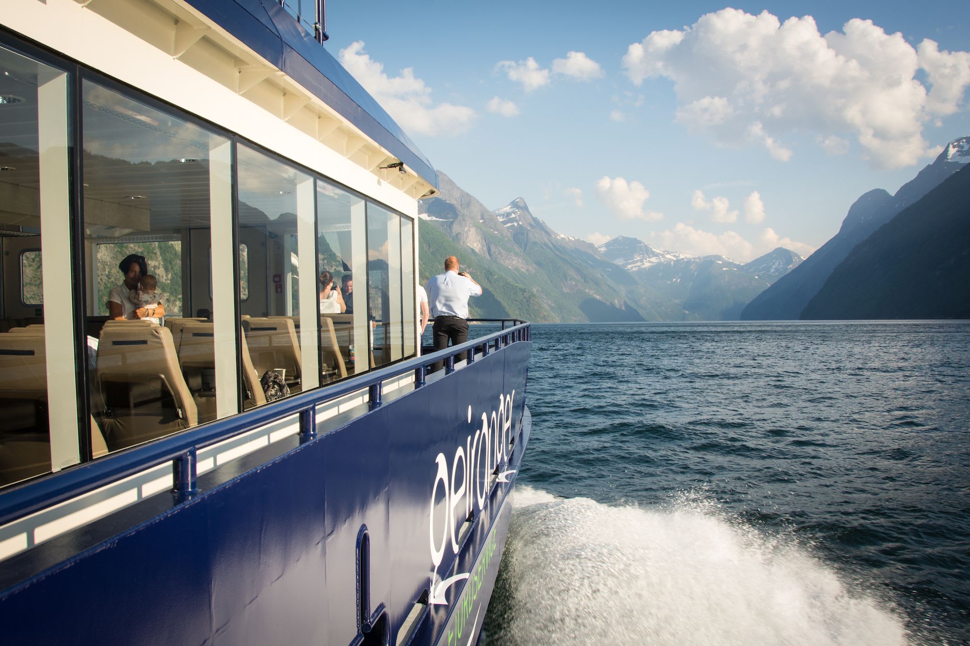 View from boat through fjord with person on deck; others enjoy scenery inside.