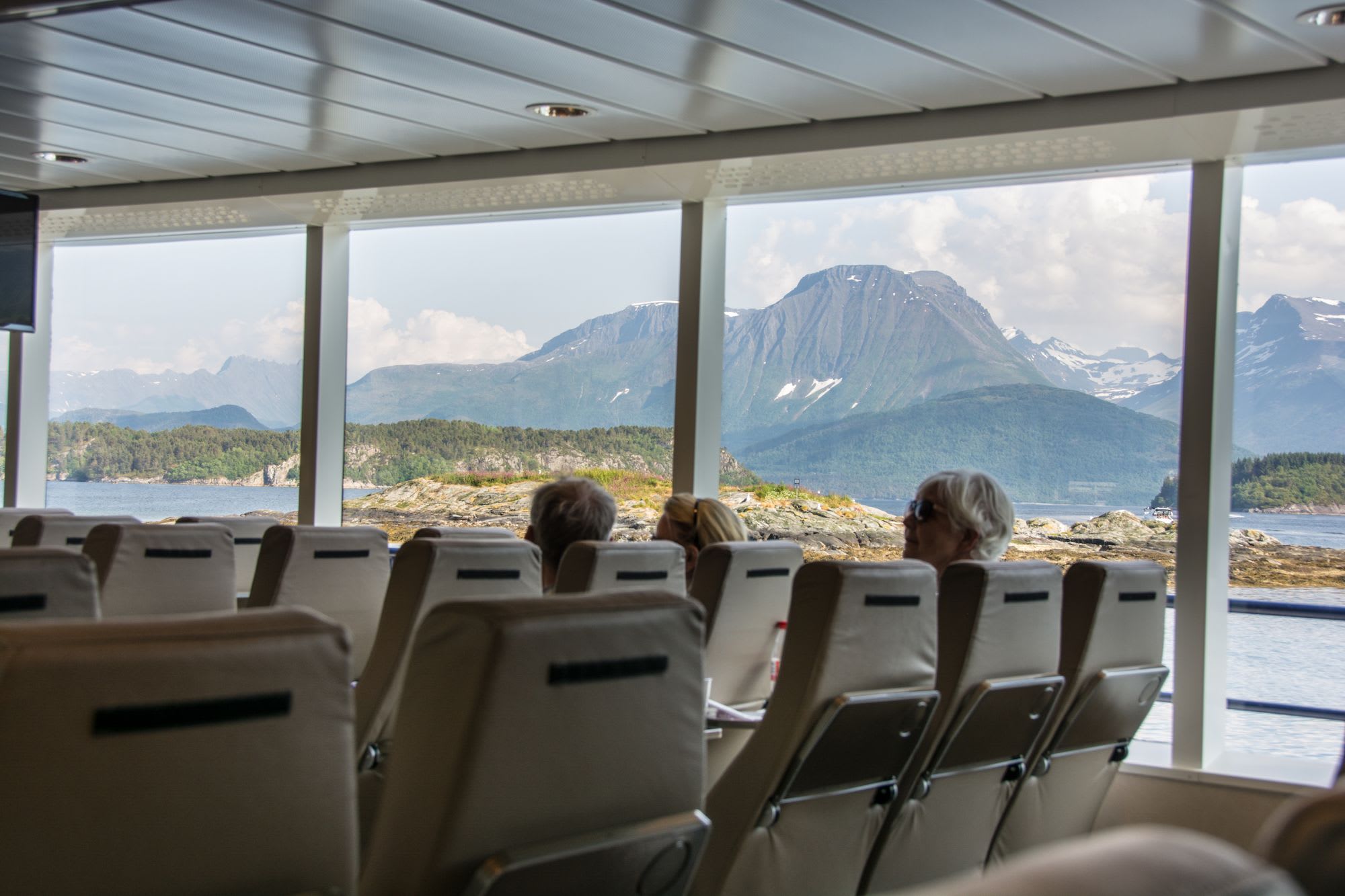 Passengers view mountains through large windows inside a sunny boat cabin.