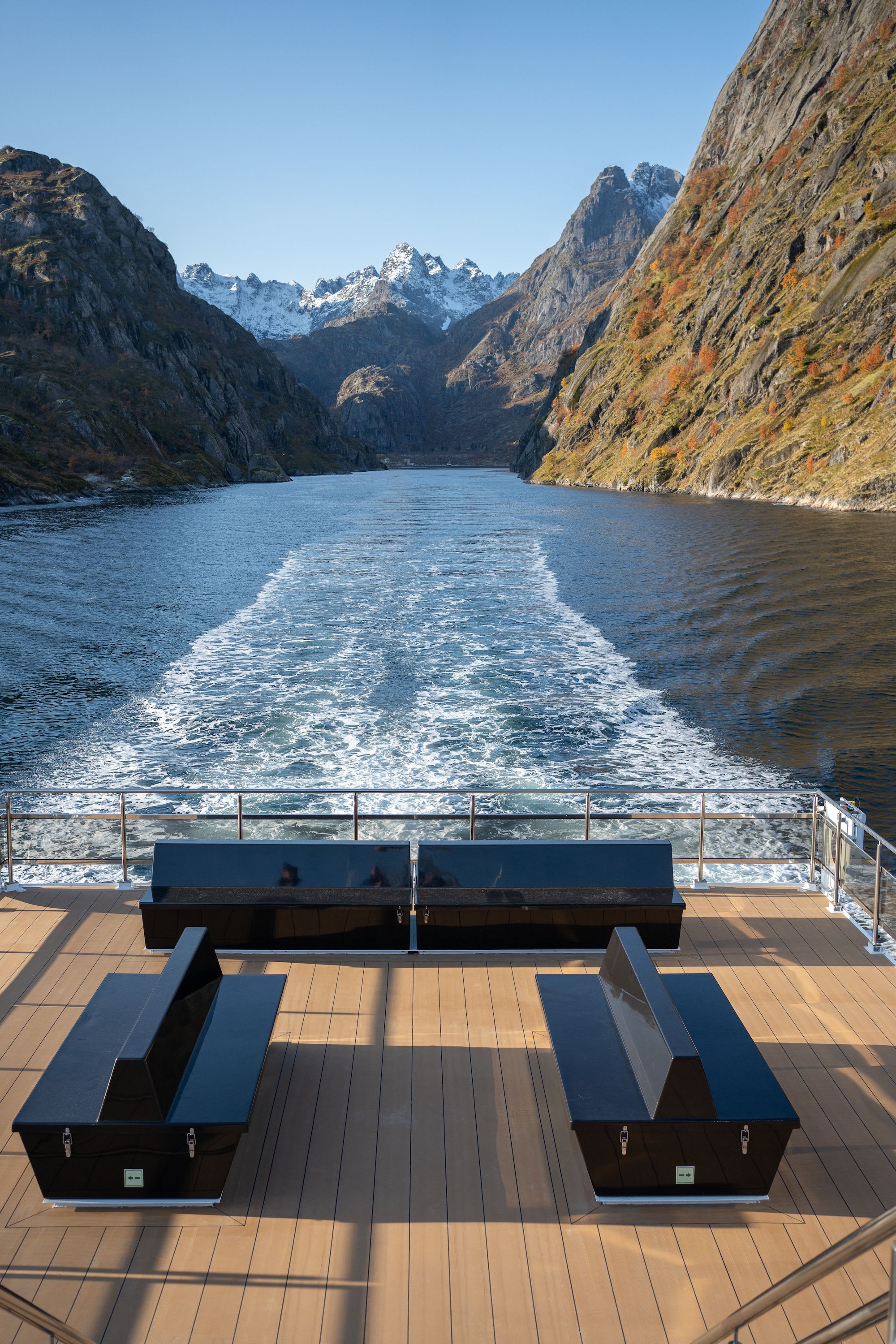 Boat wake cuts fjord between cliffs and autumn foliage.