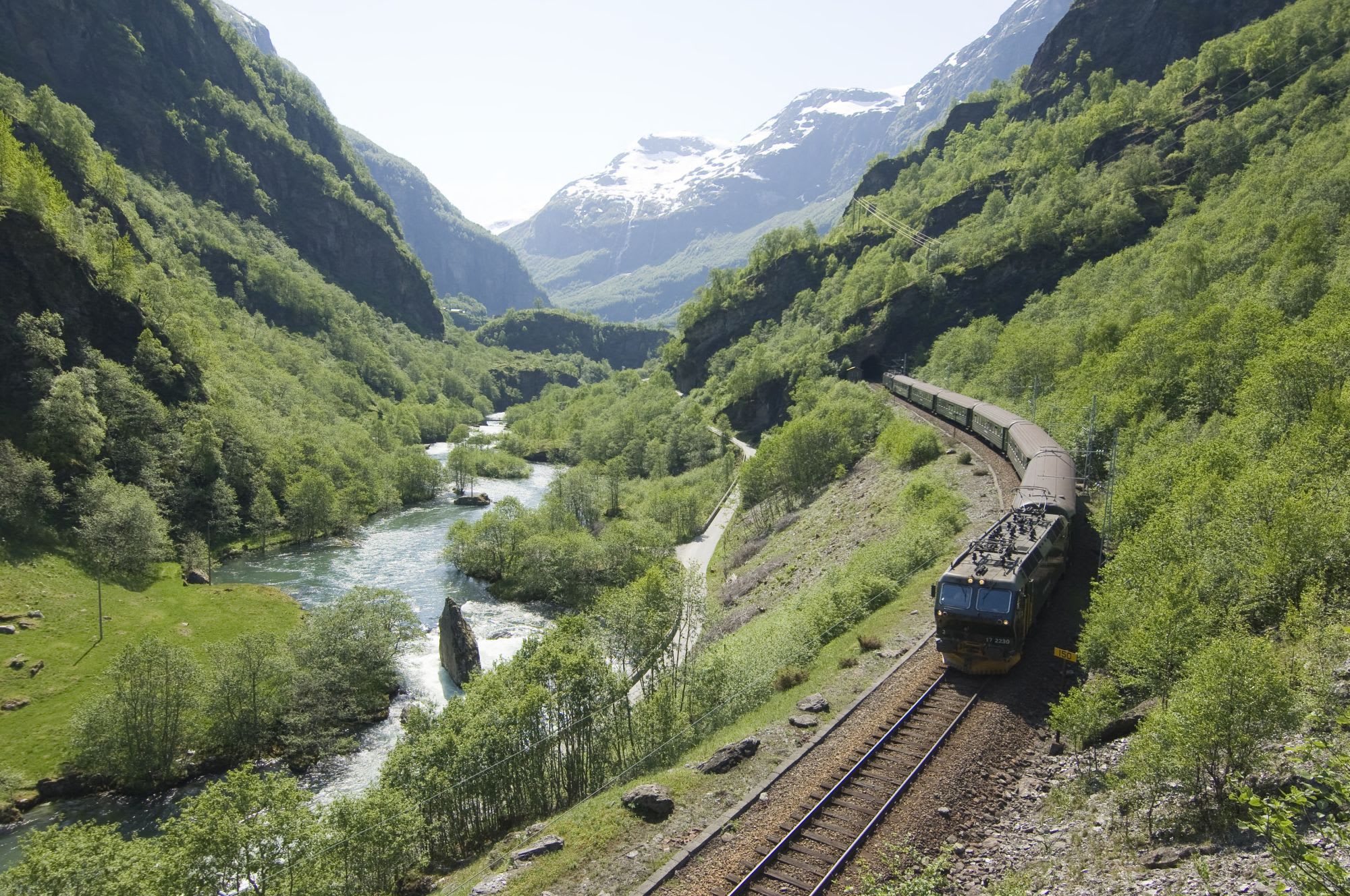 Flåmsbana kjører forbi grønne daler, brusende elver med snødekte fjell i bakgrunnen.