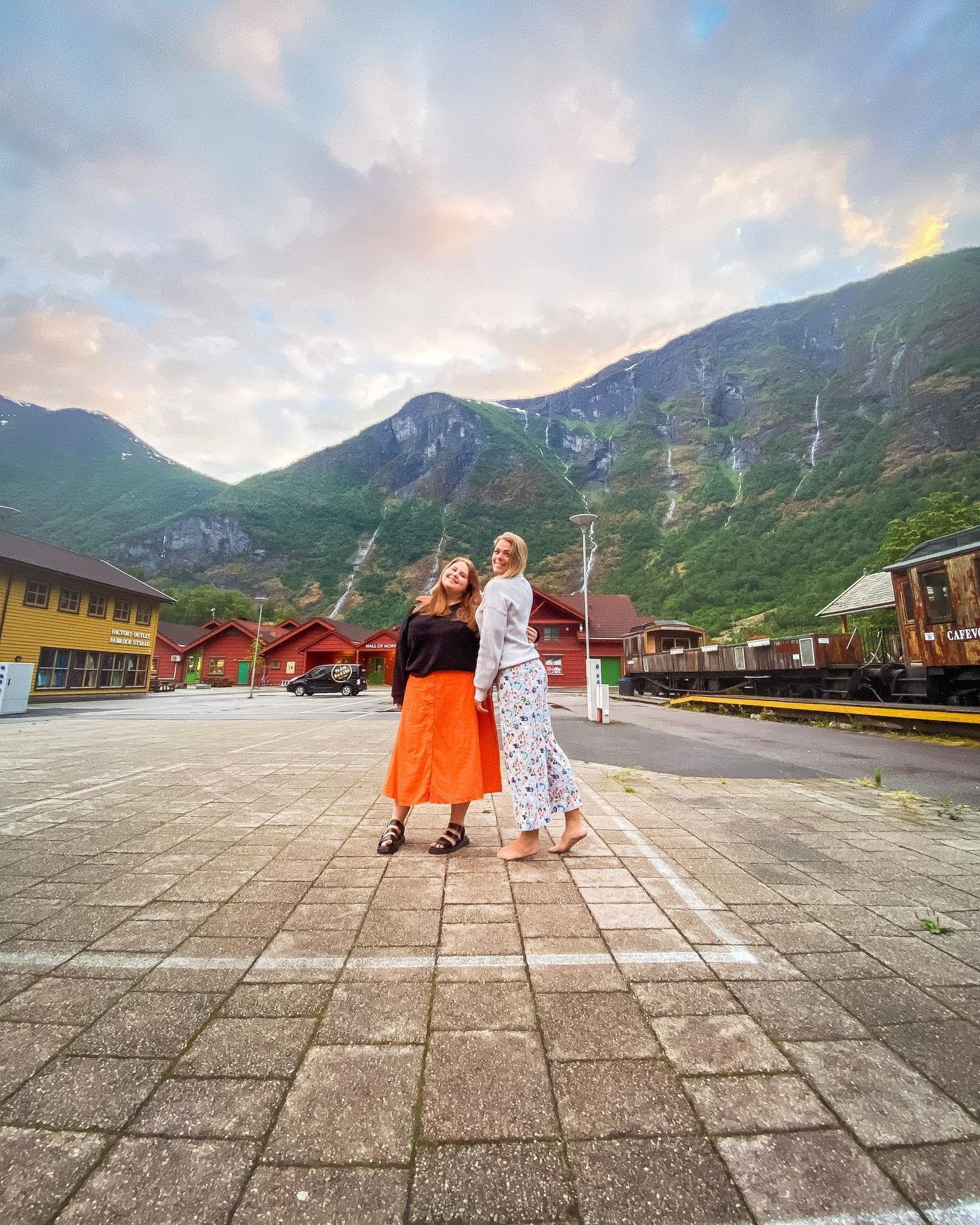To jenter som står sammen og smiler i den naturskjønne bygda Flåm med grønne fjell i bakgrunnen