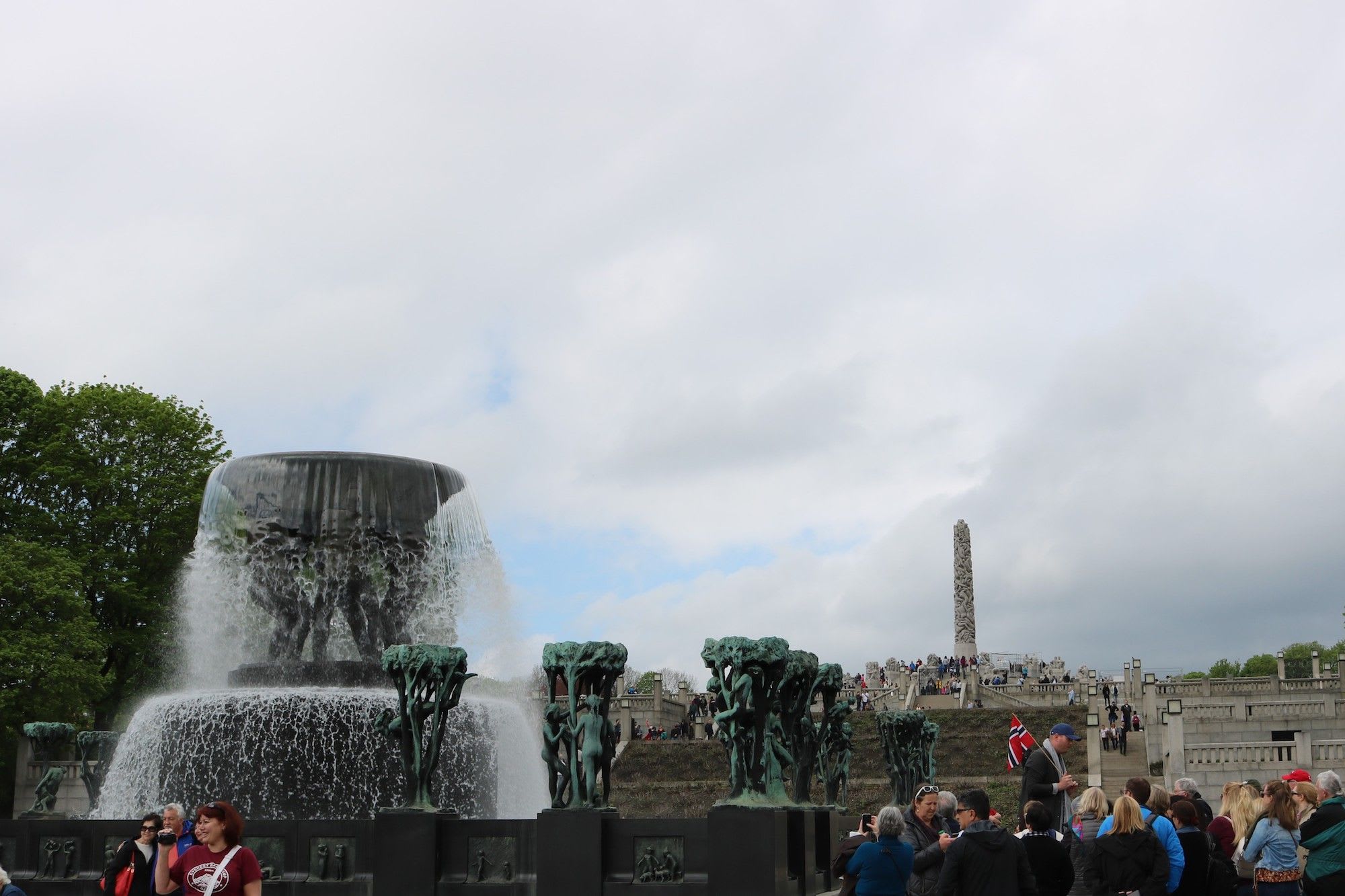 Fontene og skulpturer i park, monolitten i bakgrunnen og folkemengde med flagg.