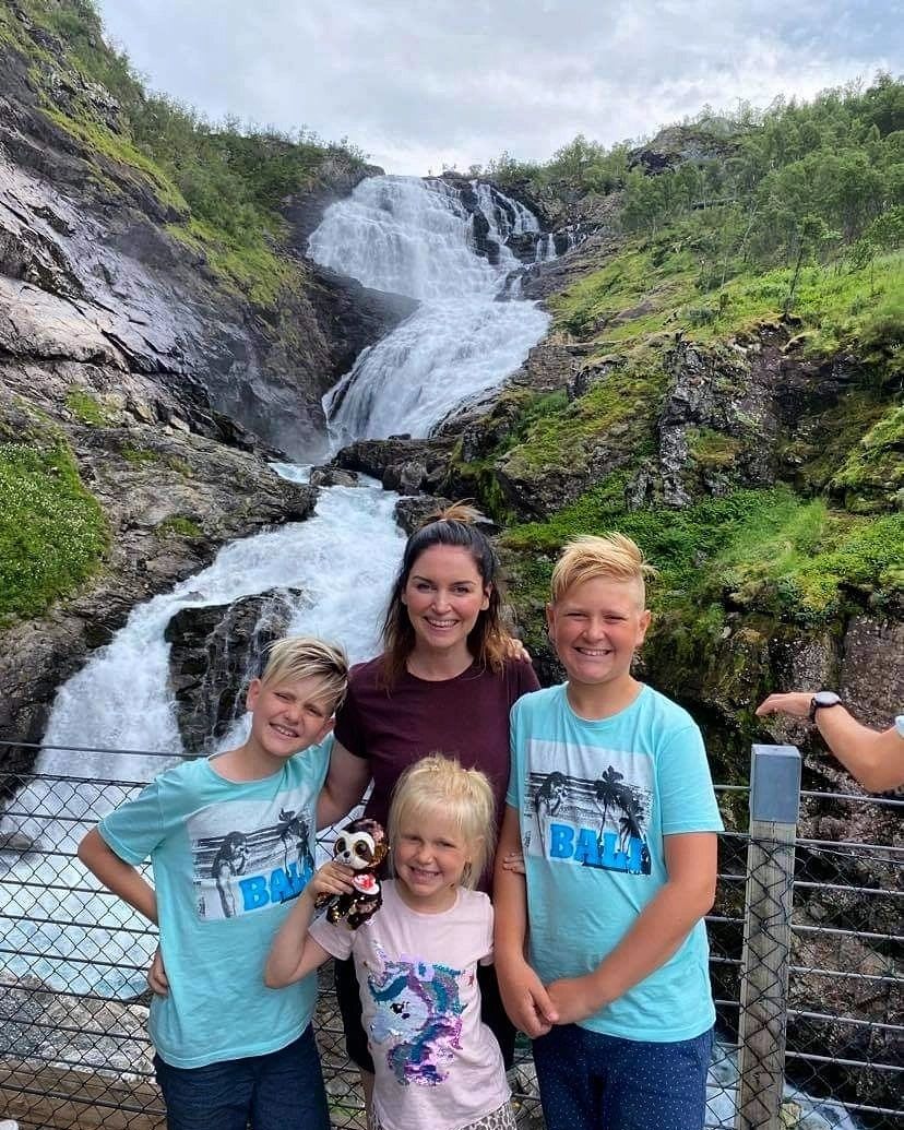 Frau und Kinder lächeln vor Kjosfossen  Wasserfall, grüne Hügel im Hintergrund.