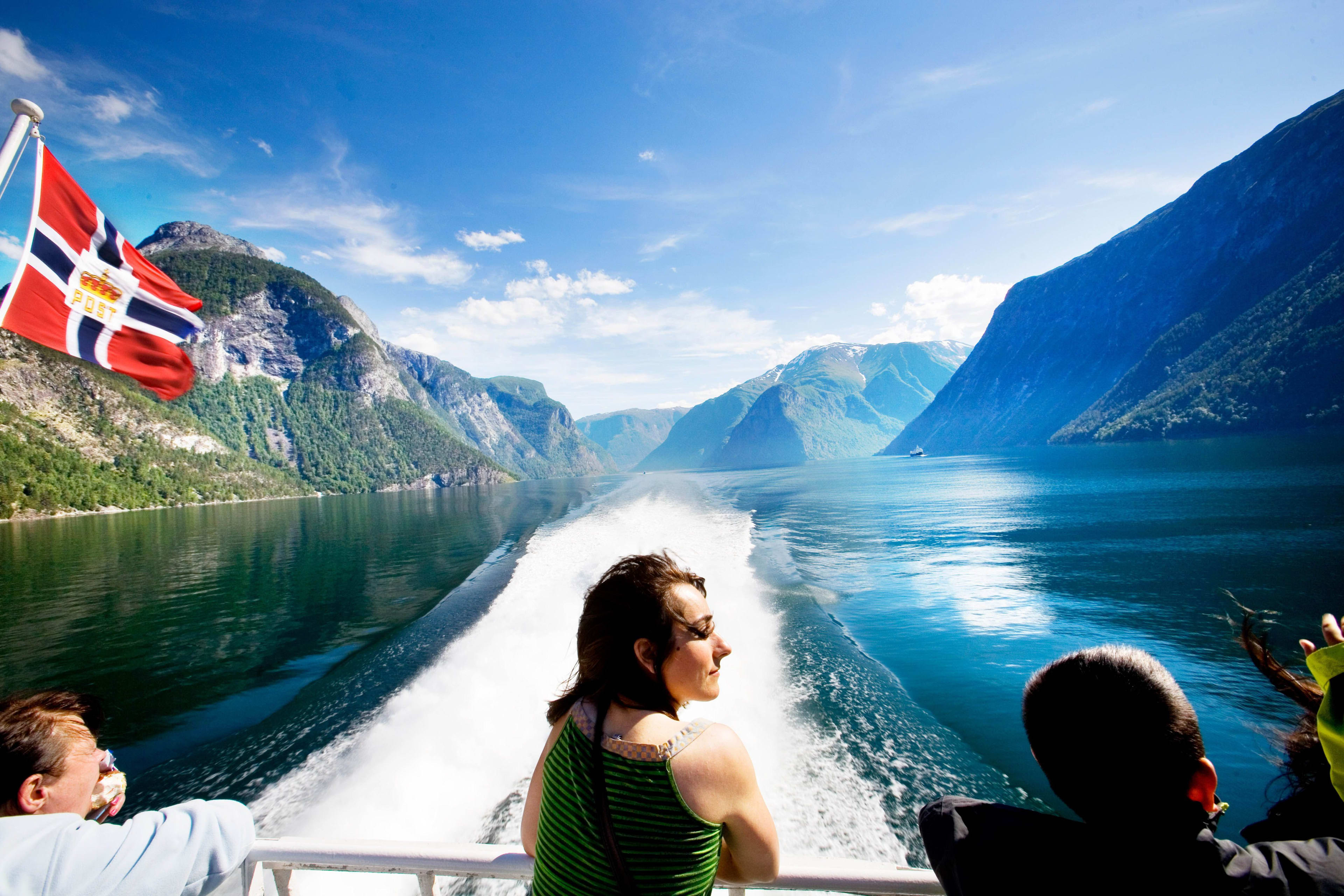 Boat on Sognefjord with flag flying, wake trailing behind on calm water.