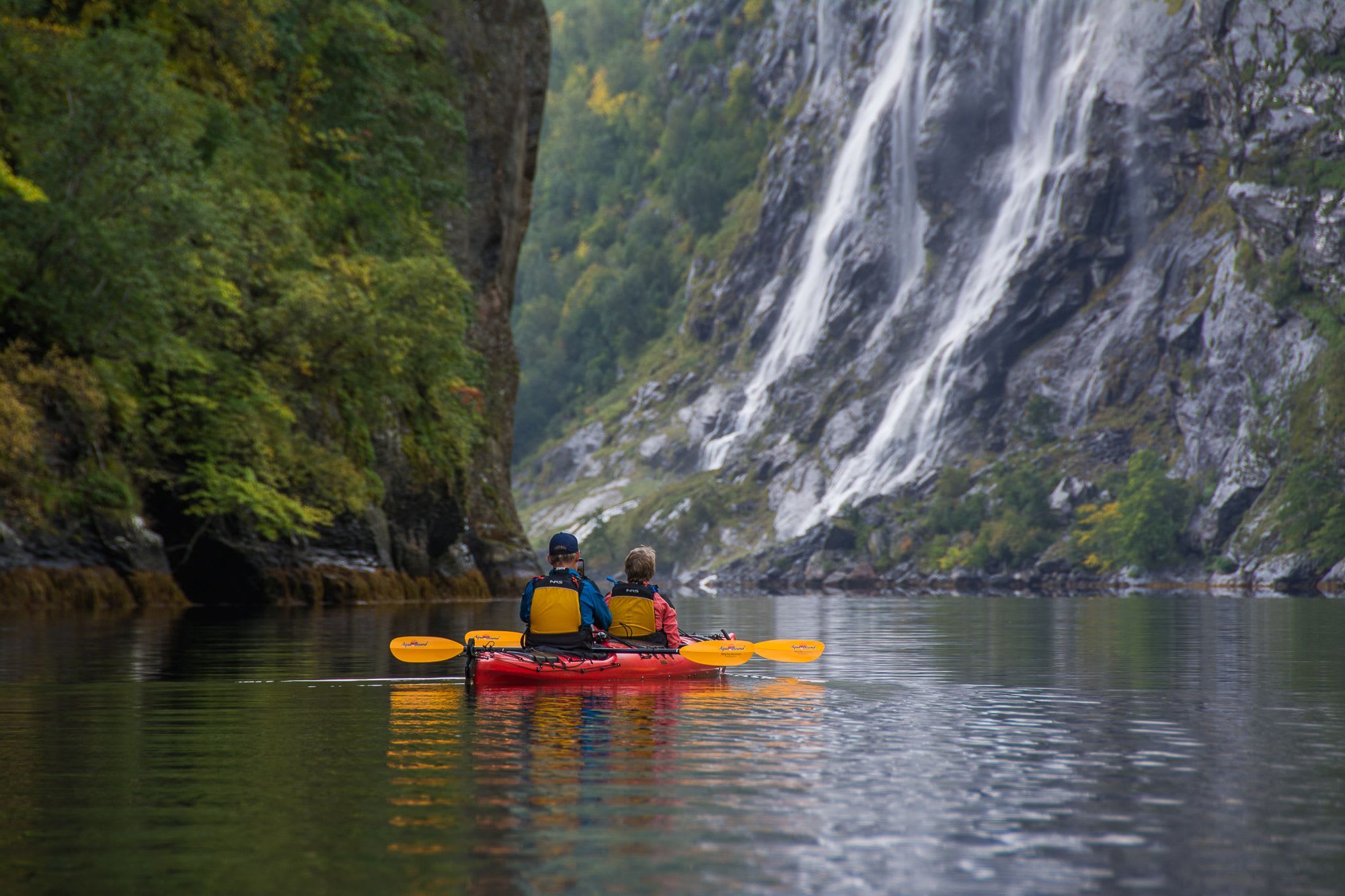 Two people in kayak near fjord waterfall.