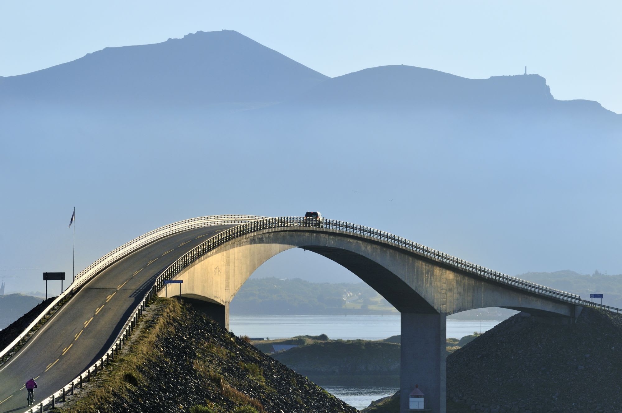 Curving bridge over water with car crossing, cyclist approaching.