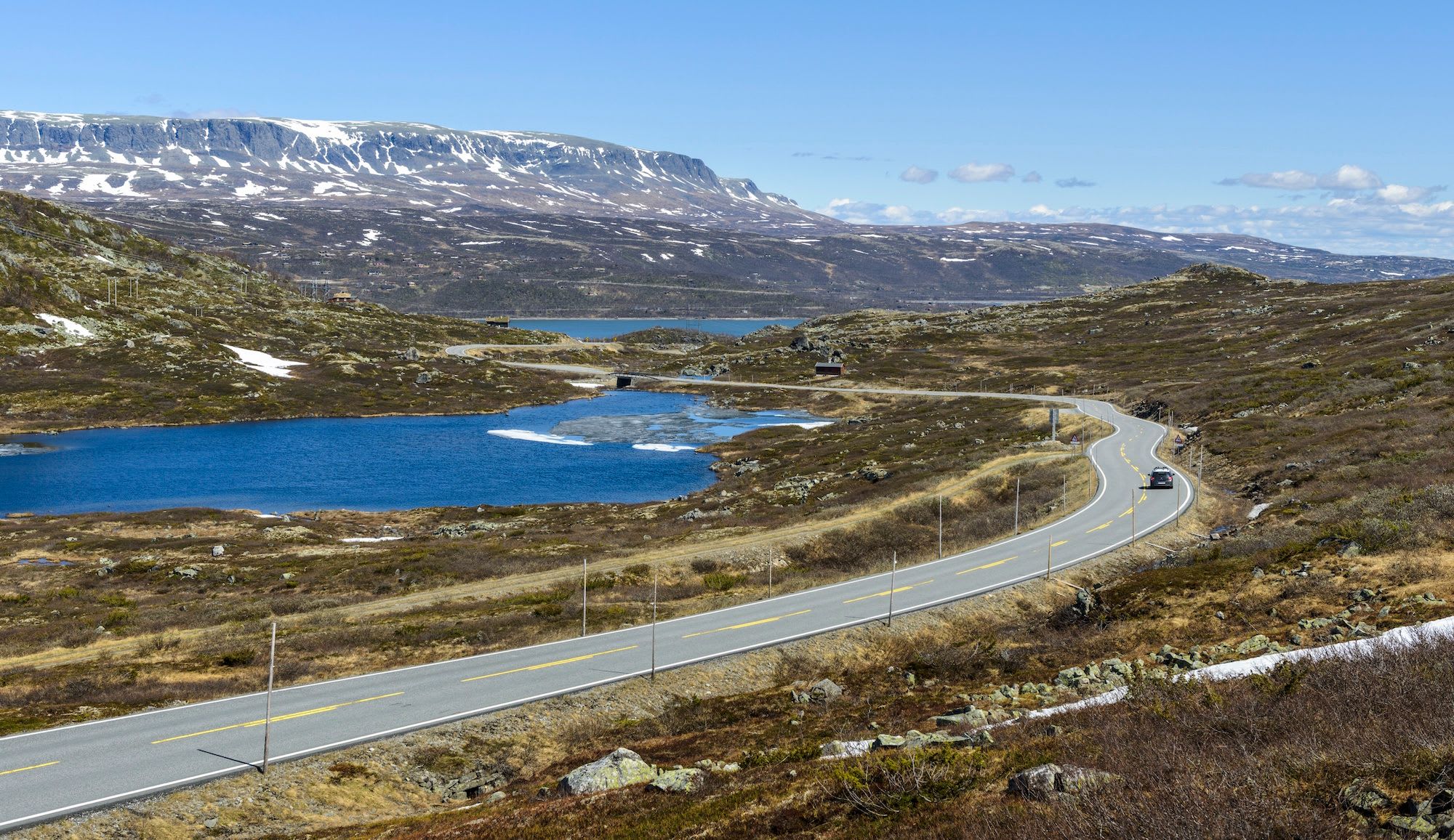 Winding mountain road by frozen lake with snowy peaks under blue sky.
