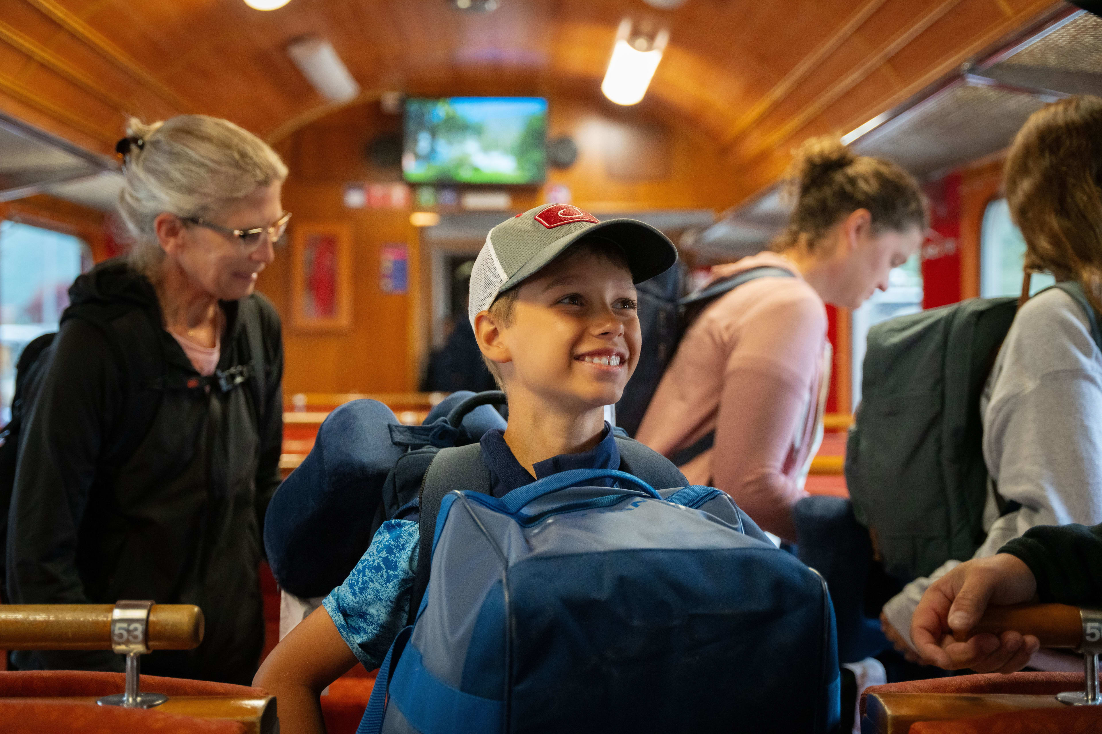 Boy with cap and big blue backpack smiles inside warm wooden train car with family.