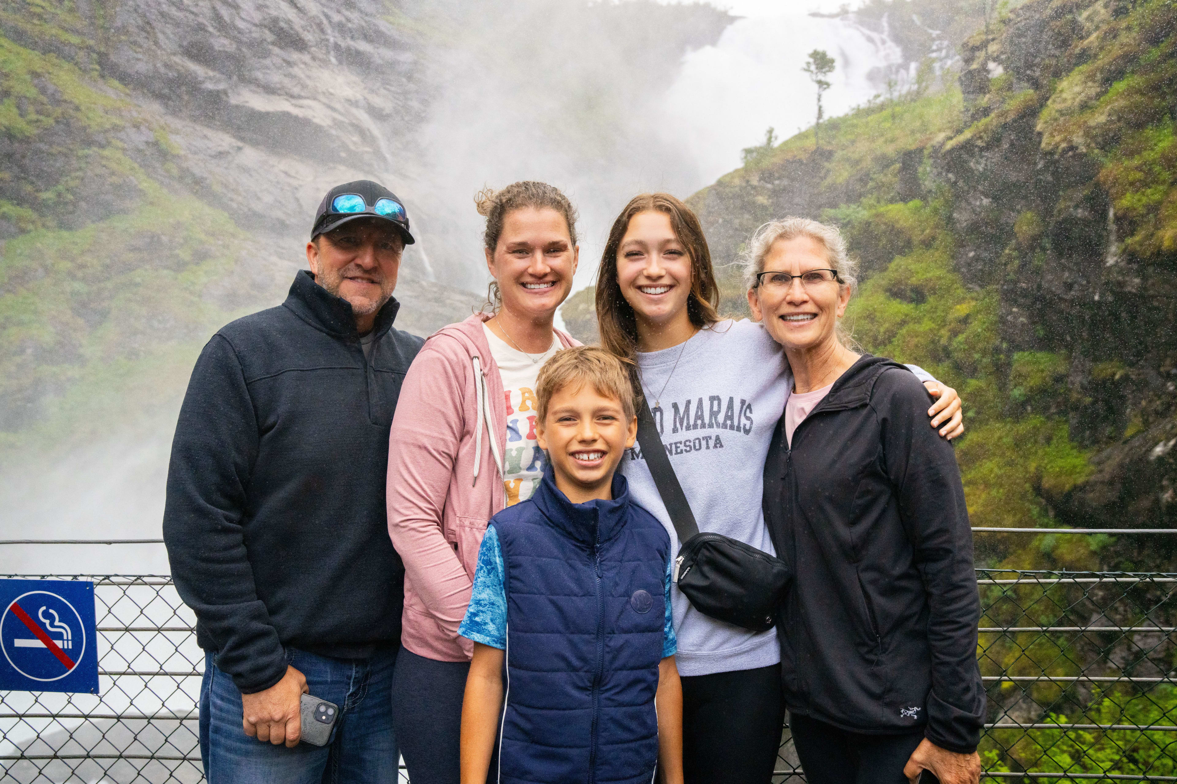 Familie på seks poserer ved foss og bratte, grønne fjell, røykeforbudsskilt synlig.