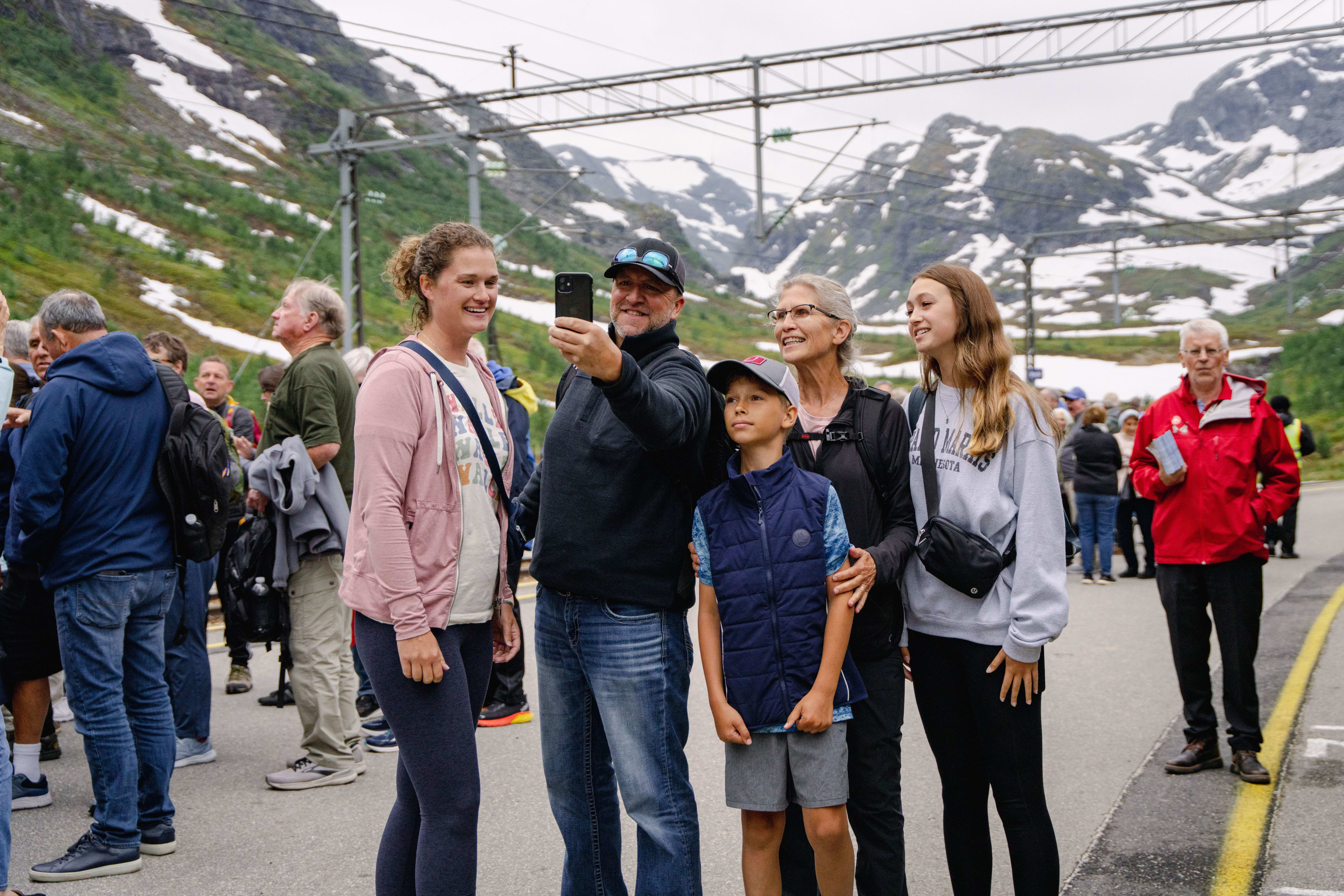 Smiling family takes selfie at mountain station with other travelers and snowy peaks.