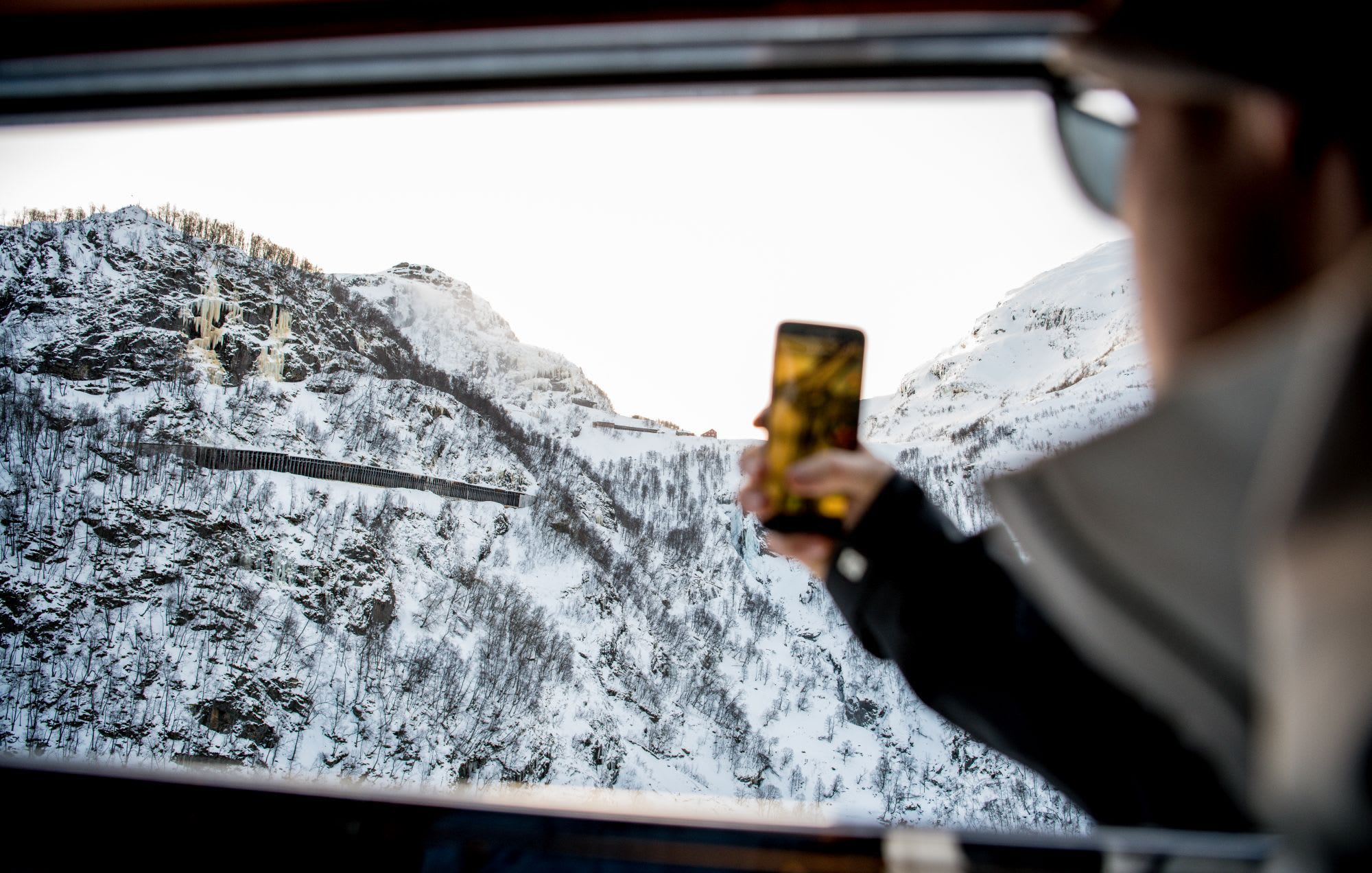 Person takes photo of snowy mountains, tunnel, and icicles from train window.