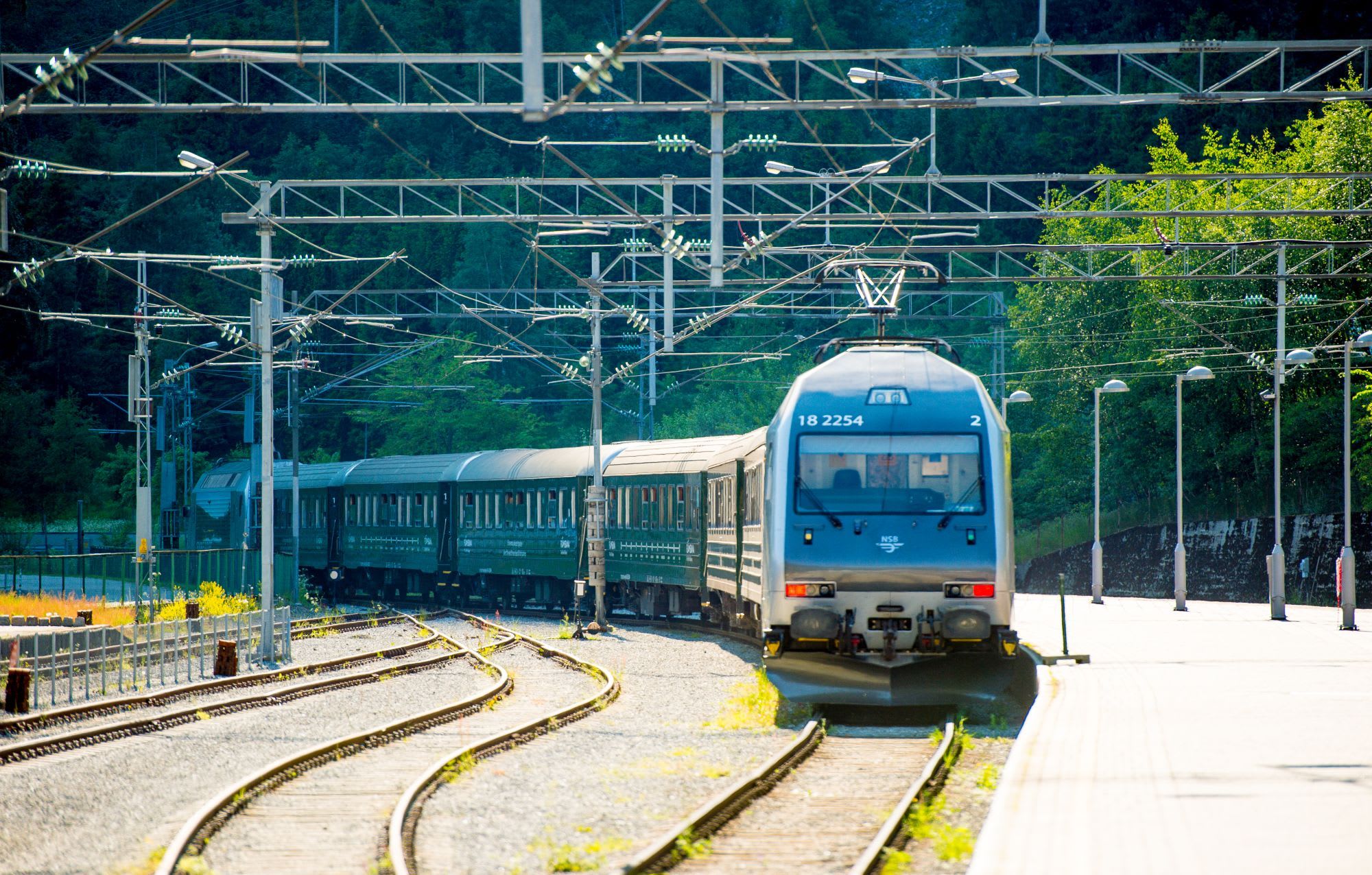 Blått lokomotiv ankommer stille stasjon omgitt av trær og ledninger.