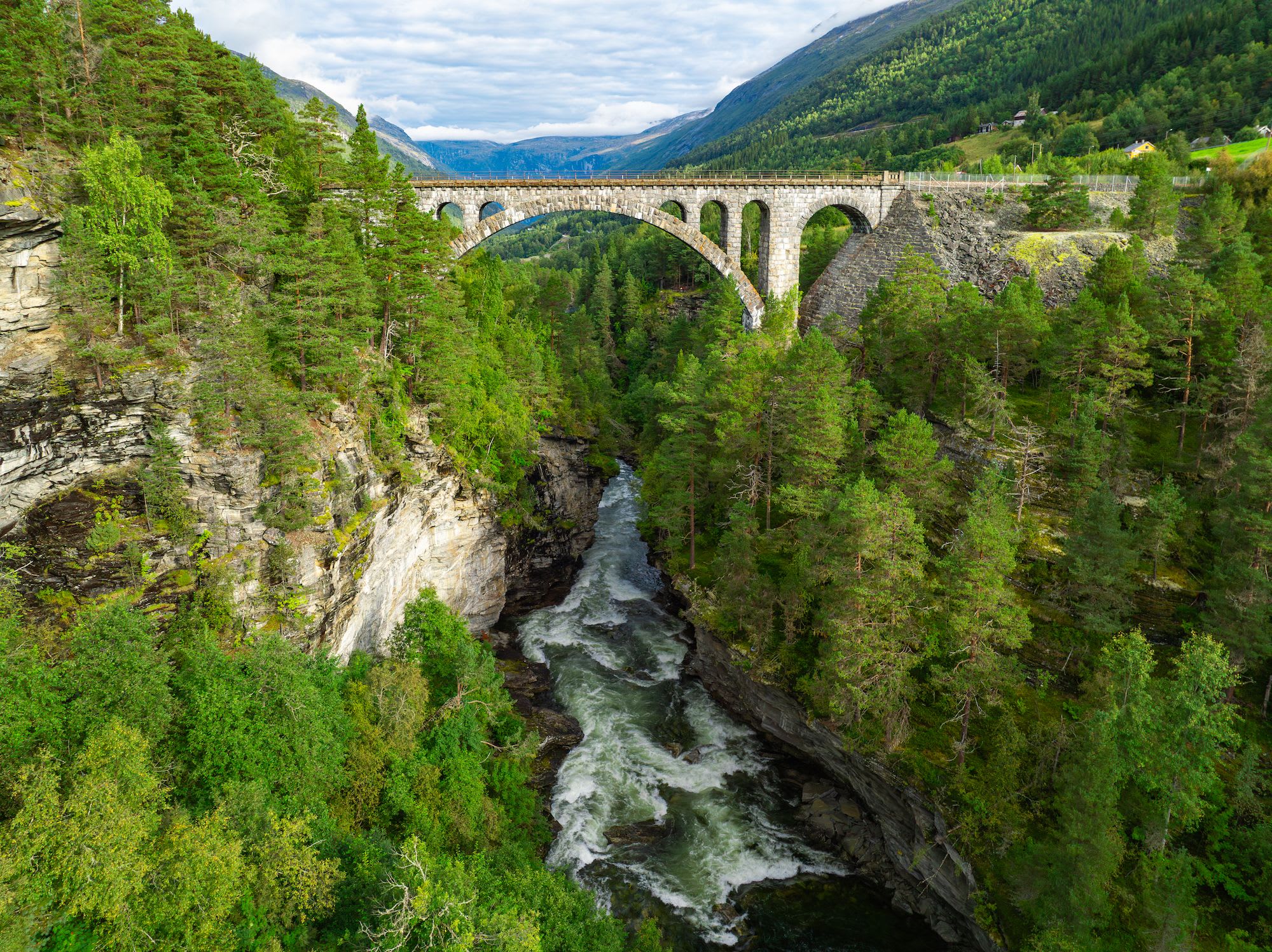 En steinbro som strekker seg over en elv med en frodig grønn skog og bratte klipper på begge sider, som fanger majesteten i naturen.