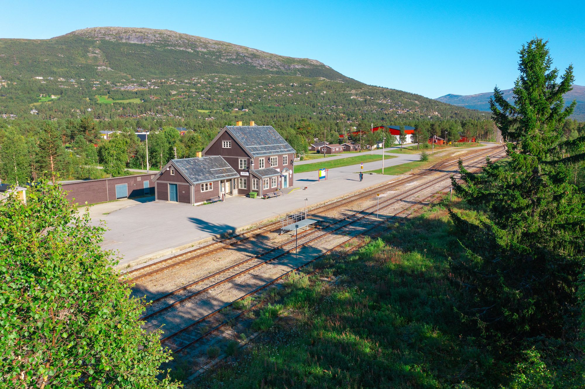 En naturskjønn togstasjon med en godt vedlikeholdt bygning, omgitt av fjell, trær og bolighus.