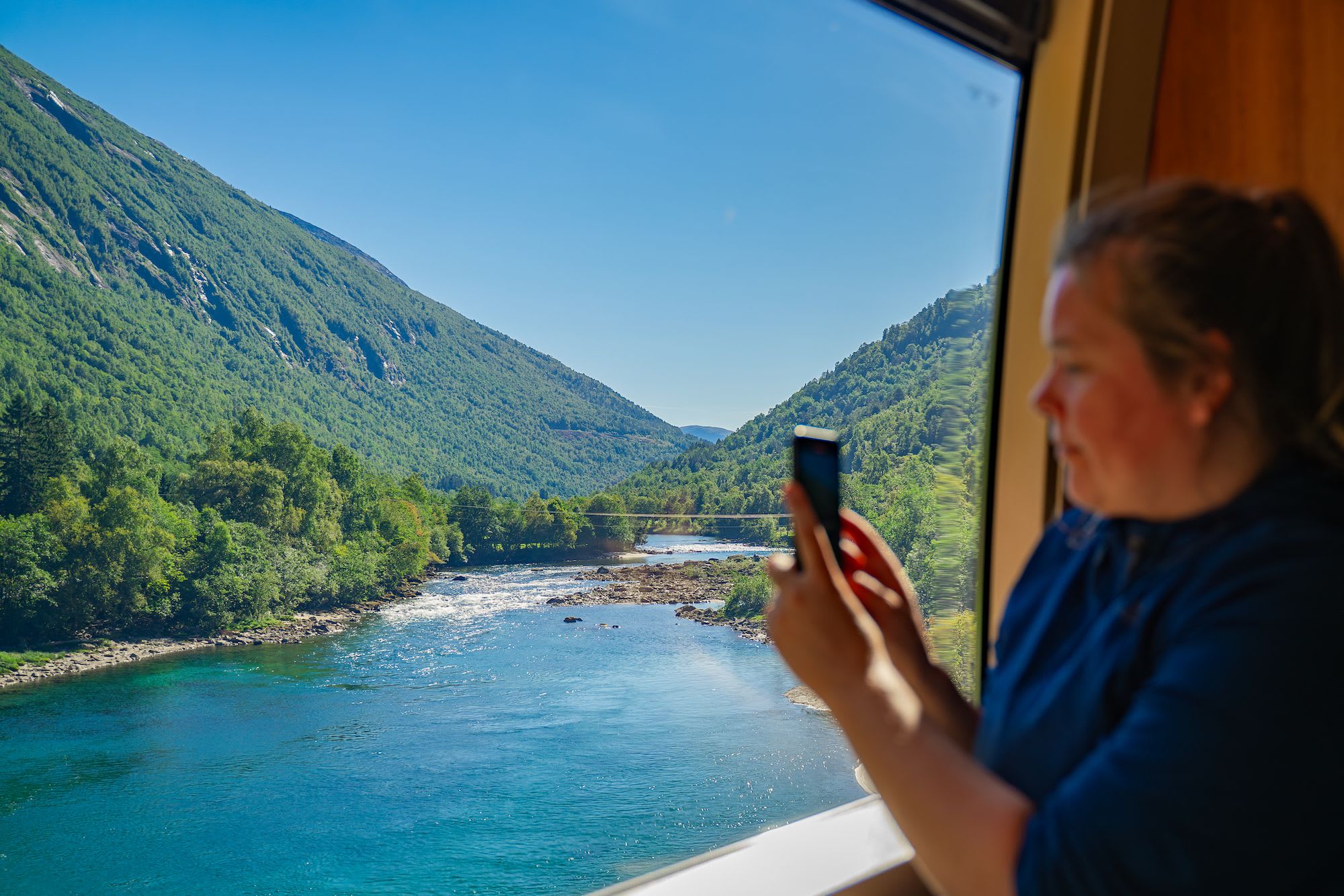 En passasjer som sitter nær vinduet i et tog, og tar et bilde av det vakre landskapet med fjell og elv.