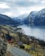 Calm fjord with houses, bare trees, and snowy mountains reflecting.