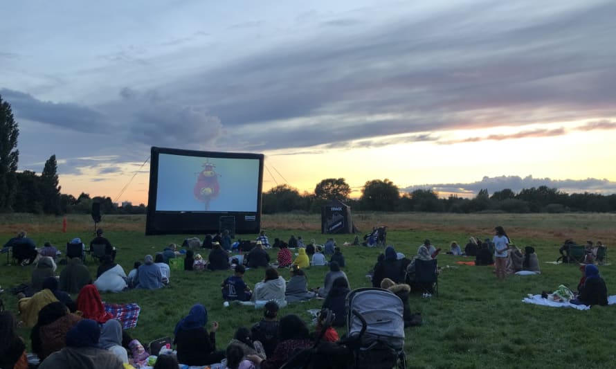 Families sitting on picnic blankets in a park watching a film on a big screen as the sun sets.