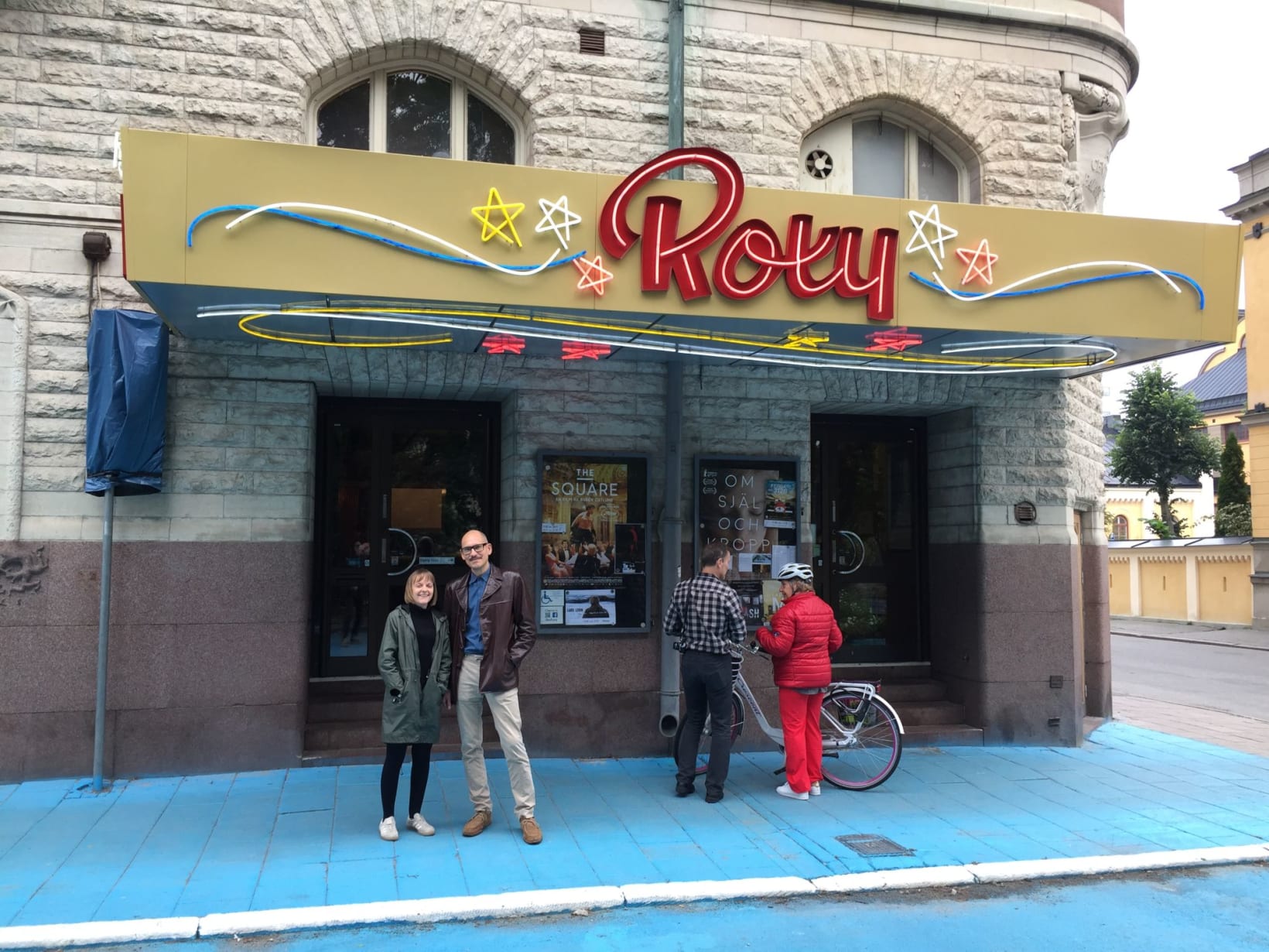 Photograph of Amy and Bo stood outside the front of the Roxy Cinema, which has an illuminated sign with stars and Roxy in large red letters. 