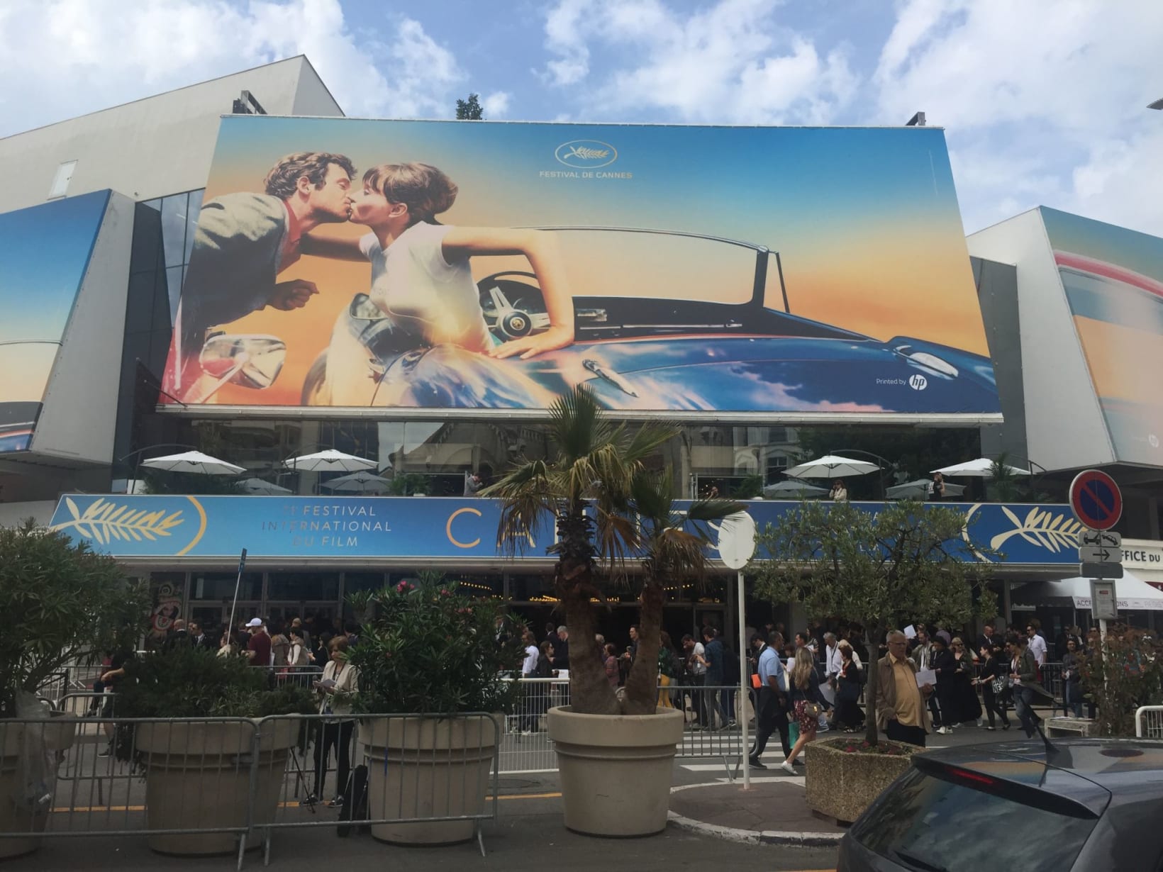 Photograph of the main entrance of the Palais des Festival. Crowds stand outside, and there are also trees, plants and a large billboard advertising the festival.