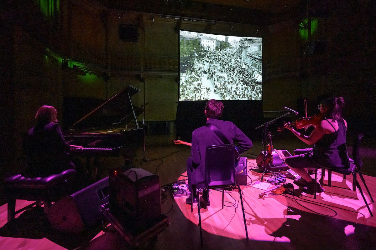 Three musicians playing piano, guitar and violin. They face away from the camera, looking at a pop up cinema screen with a black and white film playing.
