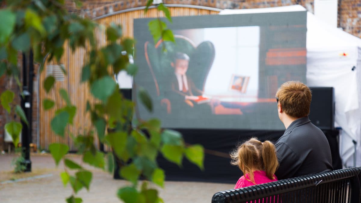 An adult and child sat on a bench watching a film on a pop up screen.