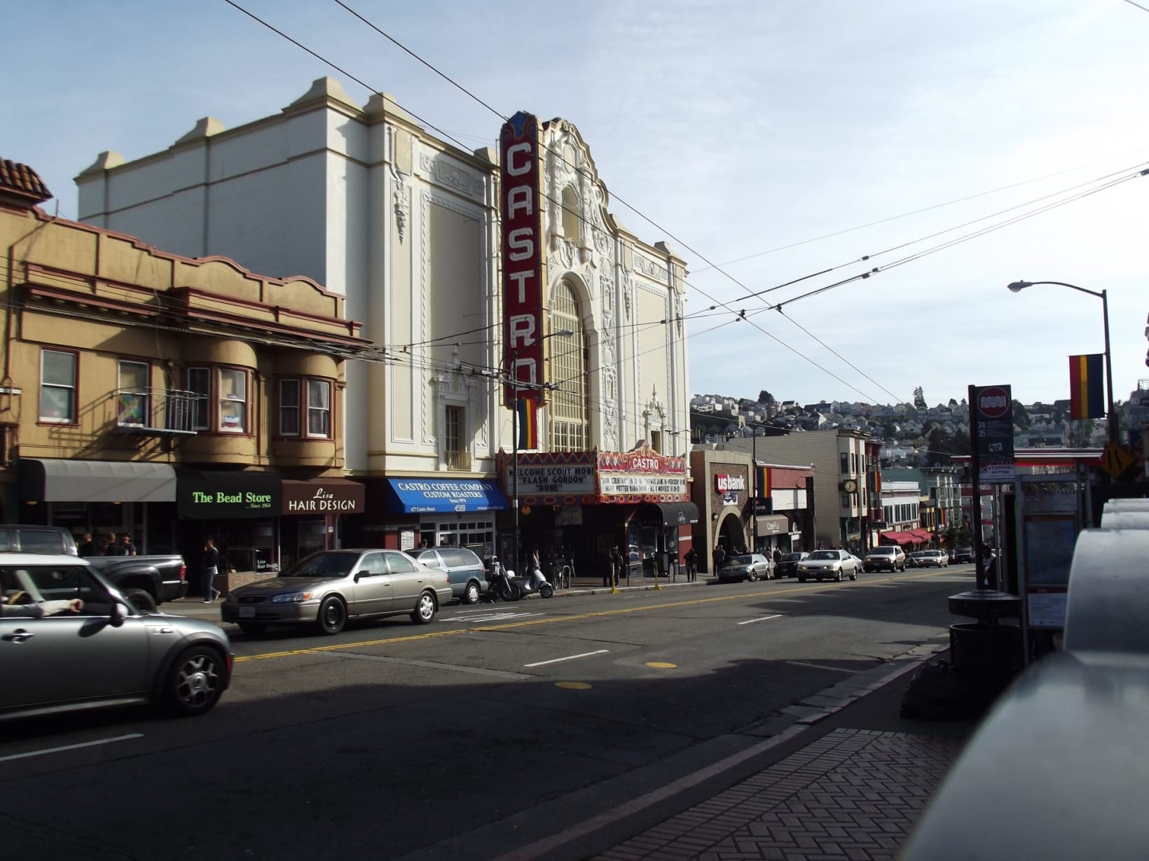 Photograph of the Castro Cinema taken from across the street. Cars are driving down the street past the cinema and other storefronts, including a bread store and a hairdressers.