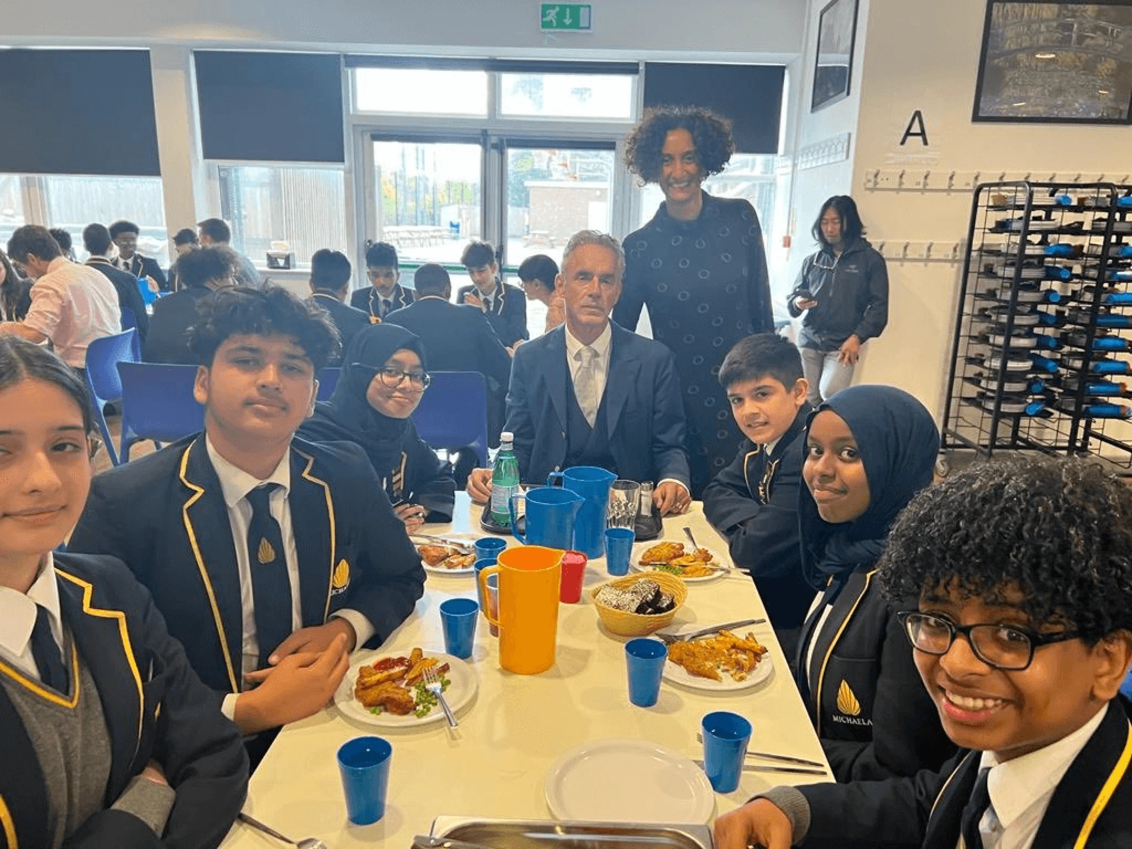 Jordan Peterson sits down with students at The Michaela Community School, whilst Katharine Birbalsingh stands behind him.