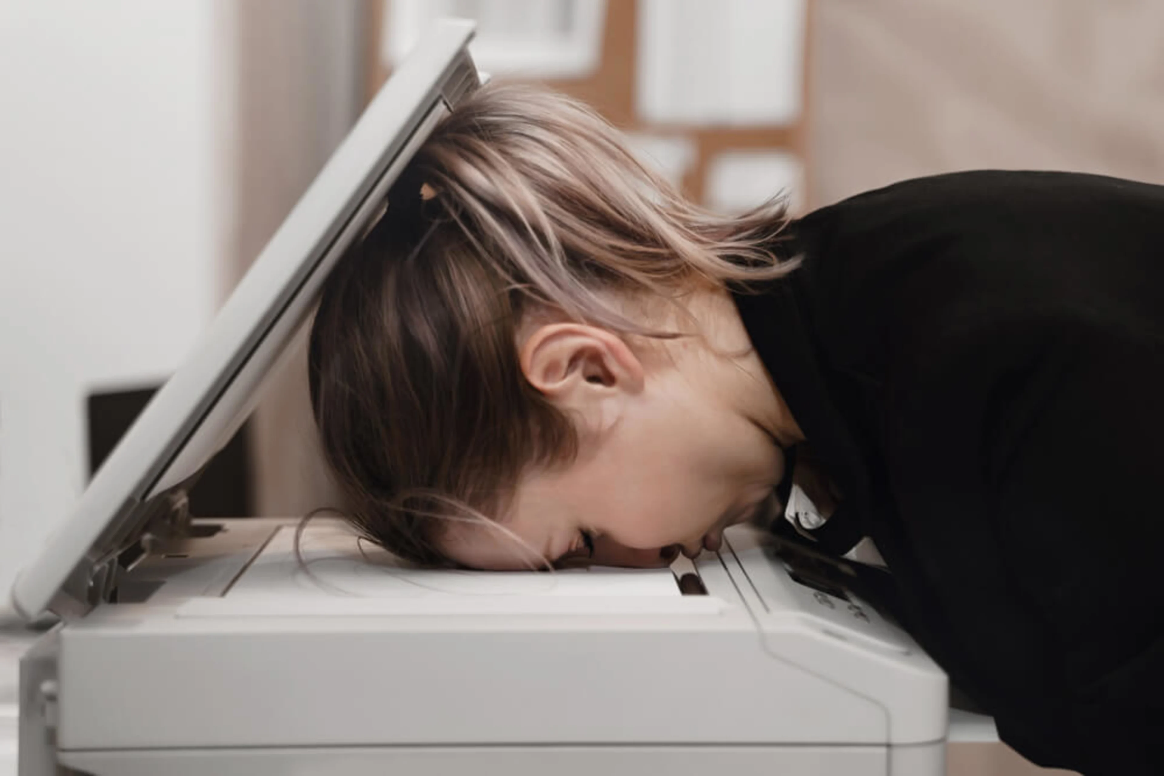 A woman resting her forehead on top of a photocopier, inside an office.