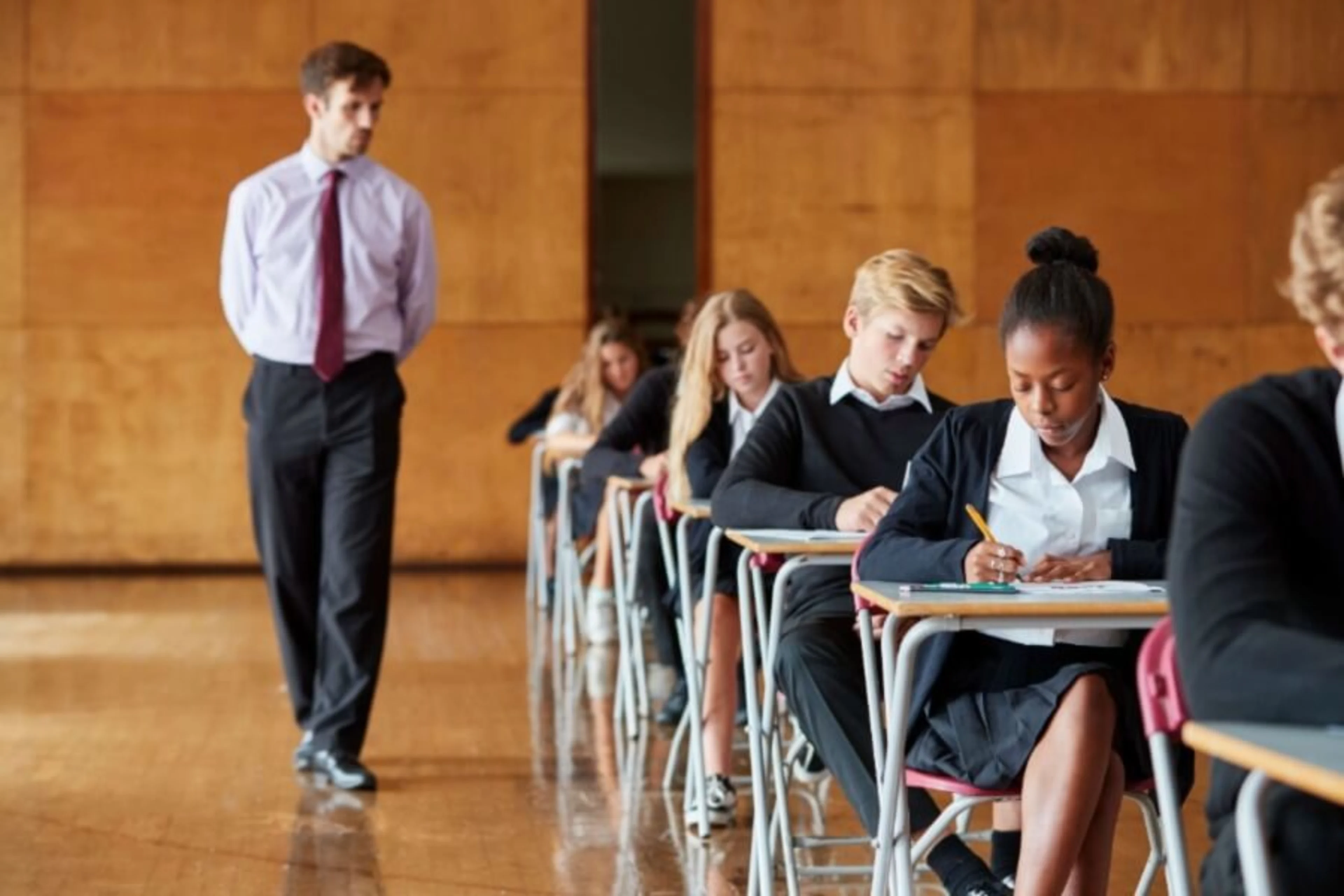 School children sit in an exam hall whilst an exam invigilator walks by