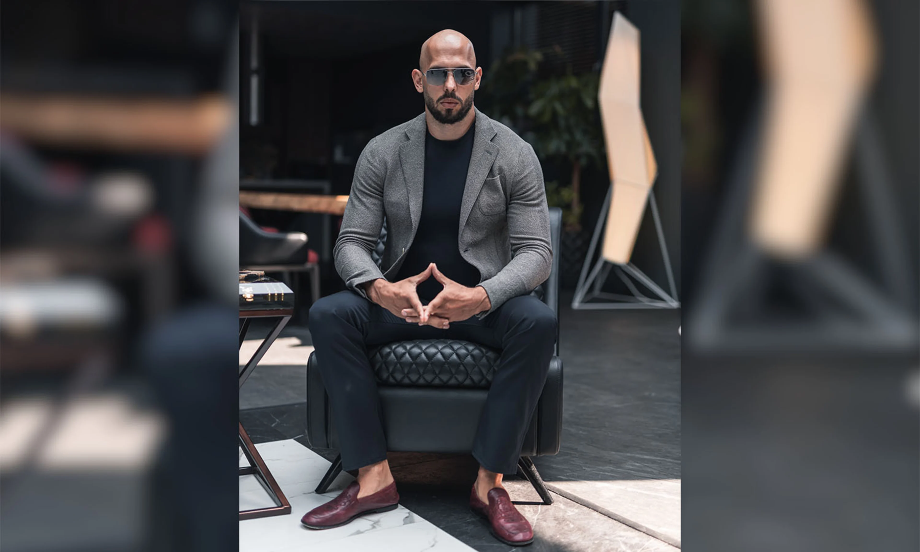 Andrew Tate posing in a chair with his distinct scholar's cradle, wearing sunglasses