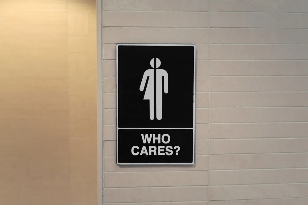 A black bathroom sign on a white wall, with a door to the left. The sign has an image of a person on it, half man, half woman. Beneath it are the words “Who Cares?”.