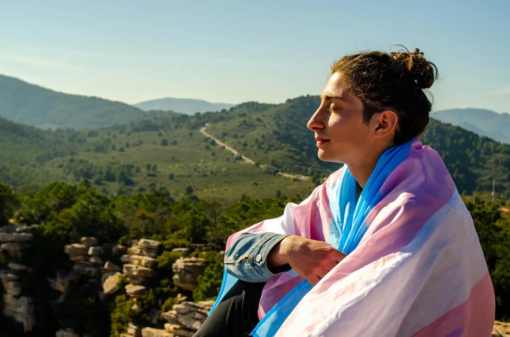 A person sitting with the trans pride flag wrapped around their shoulders, with mountains in the distance.