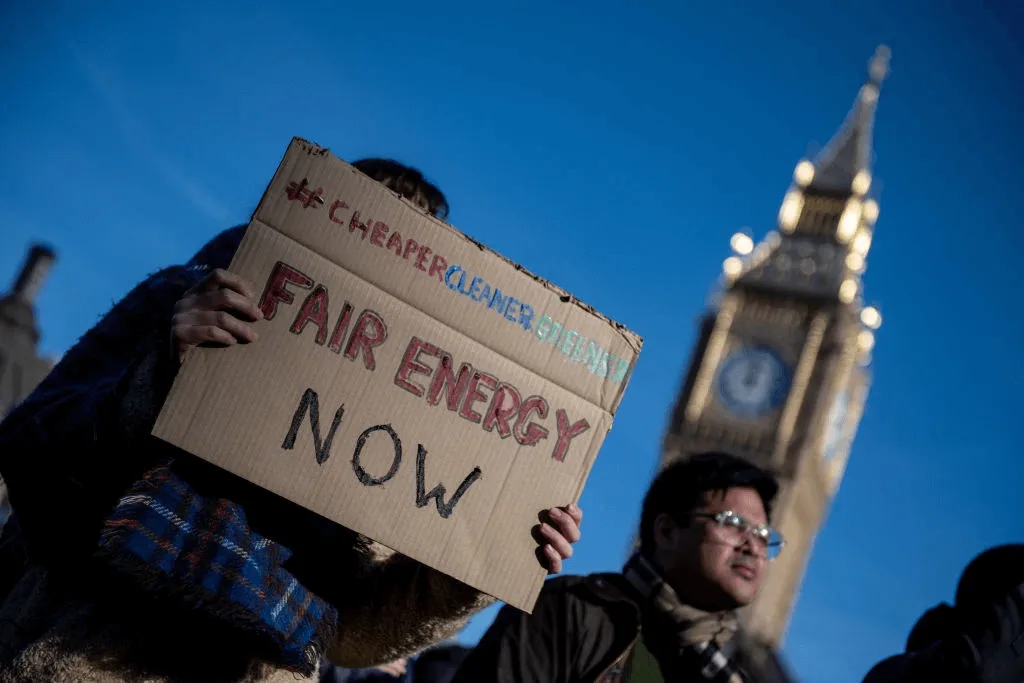 A woman holds up sign outside the palace of Westminster, with the message "Cheaper, Cleaner, Greener. Fair energy now."