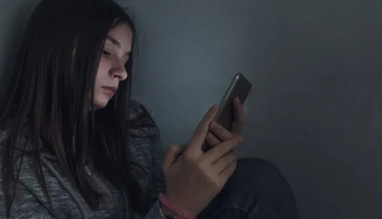 A sad teenage girl looking at her phone in a dark room.