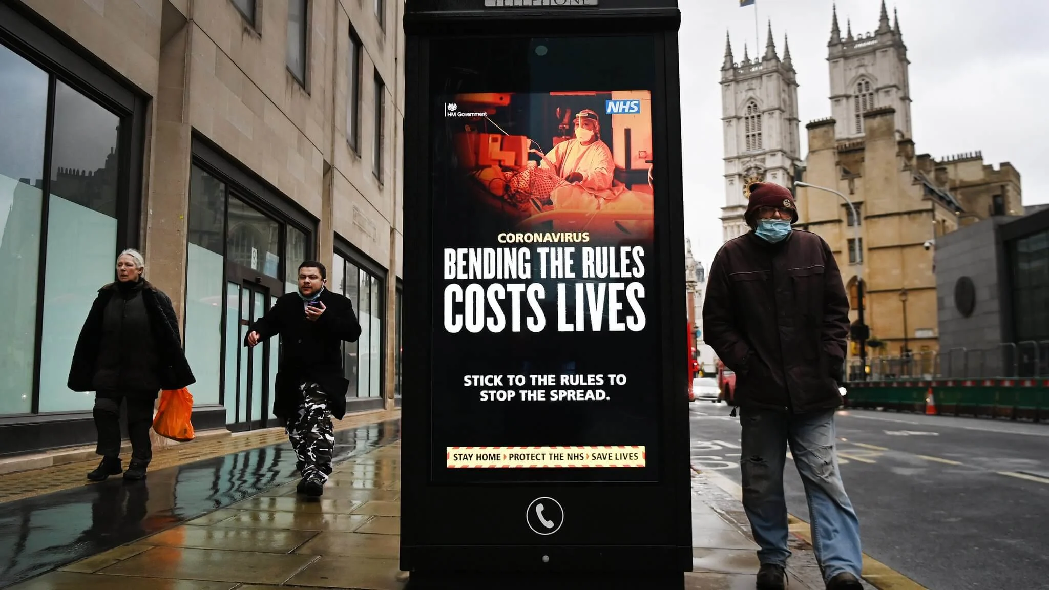 A near almost empty street in London during the COVID-19 pandemic. An elderly man wearing a face mask walks past a government-sponsored advertisement with the words "Coronavirus: bending the rules costs lives. Stick to the rules to stop the spread. Stay home. Protect the NHS. Save lives."