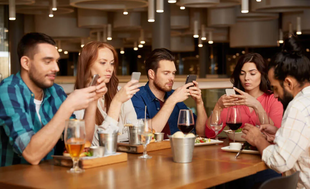 Five friends sitting around a table at a restaurant. No one is engaging in conversation. Everyone has their eyes glued to their mobile phones.