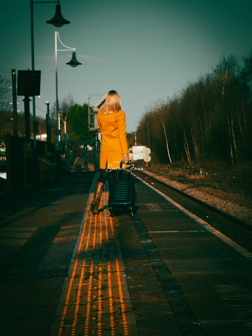 "Woman in yellow coat with blue wheeled case waiting at a train station under a blue sky." stock image